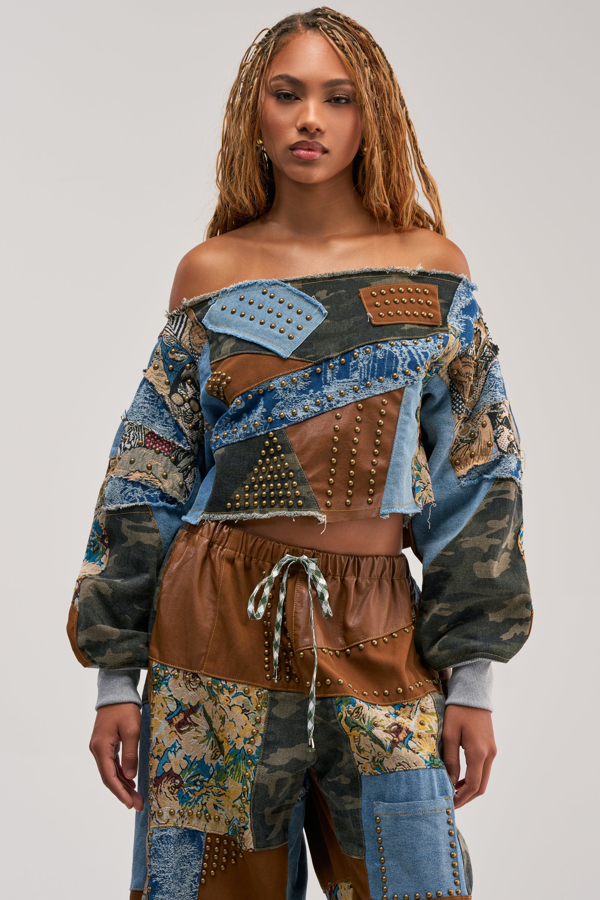 A woman with long braids wears the CALL ME CRAZY PATCHWORK OFF THE SHOULDER SWEATSHIRT IN BROWN MULTI and matching pants—featuring bold denim, leather, camo, and studded details—for a standout streetwear look against a plain gray background.