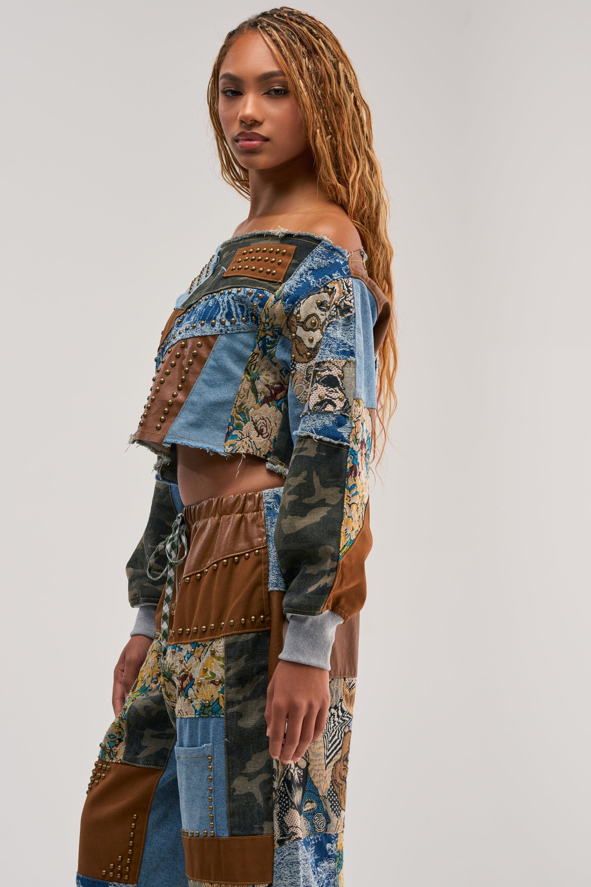 Wearing the CALL ME CRAZY PATCHWORK OFF THE SHOULDER SWEATSHIRT IN BROWN MULTI, a person with long braided hair poses sideways against a plain light gray background. The bold streetwear piece features a patchwork of denim, camo, floral fabrics, and metal studs for an eye-catching look.