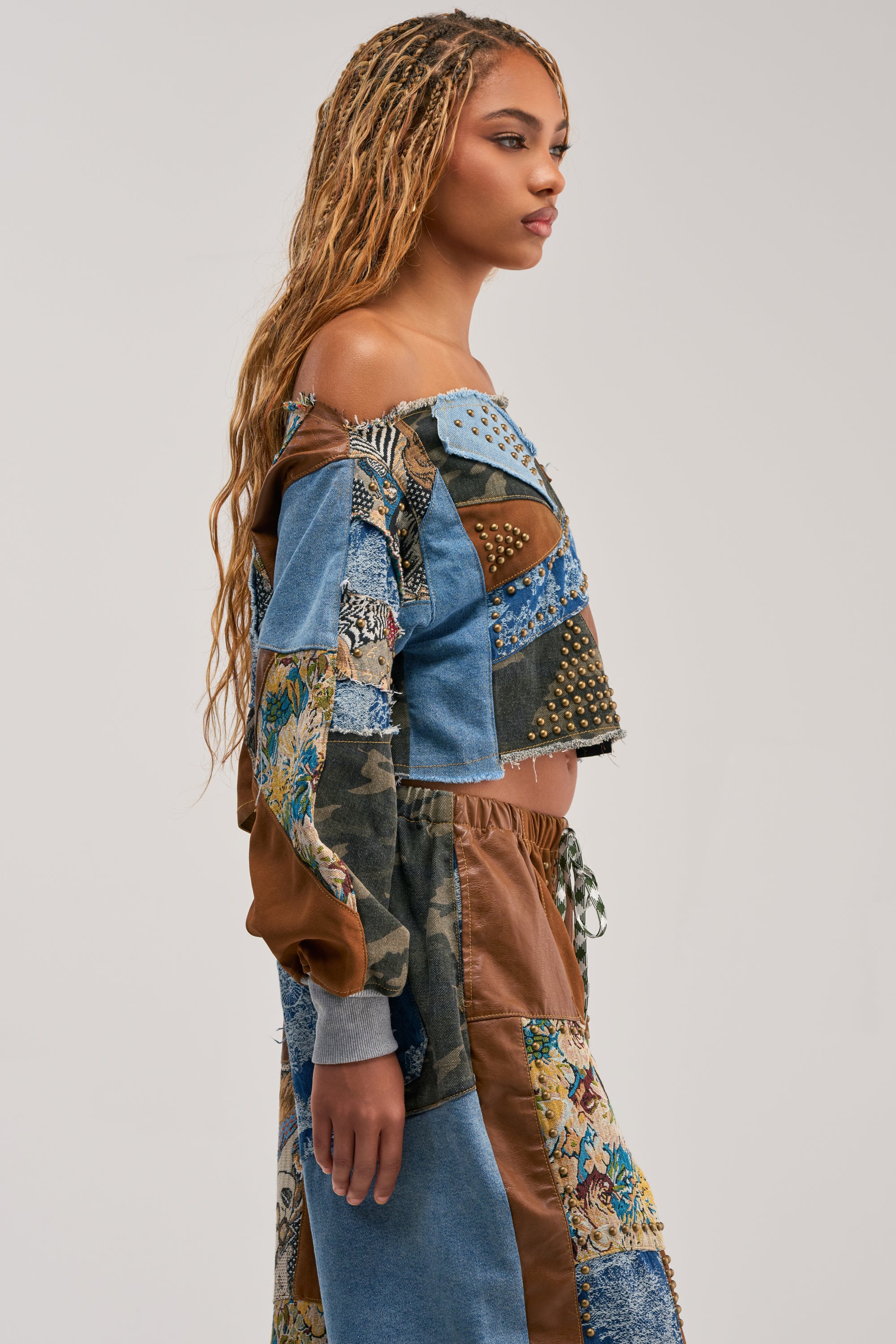 A woman with long, braided hair stands in profile against a plain background, wearing the CALL ME CRAZY PATCHWORK OFF THE SHOULDER SWEATSHIRT IN BROWN MULTI—a streetwear-inspired piece featuring patchwork denim and fabric, floral accents, and studded details—paired with a matching skirt.