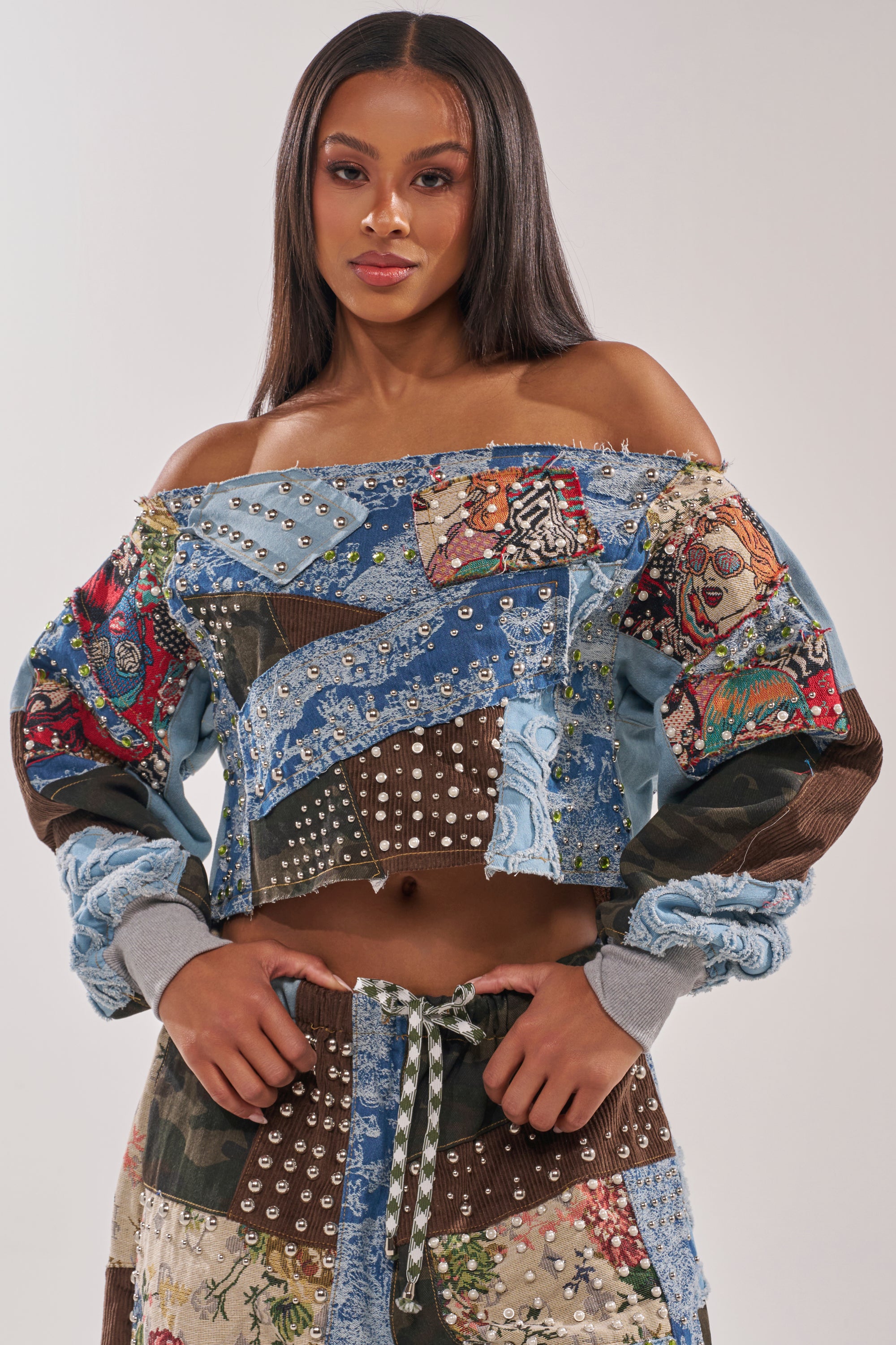 A woman with straight, long hair models the CALL ME CRAZY PATCHWORK OFF THE SHOULDER SWEATSHIRT and matching pants, embodying elevated streetwear style against a plain light background.