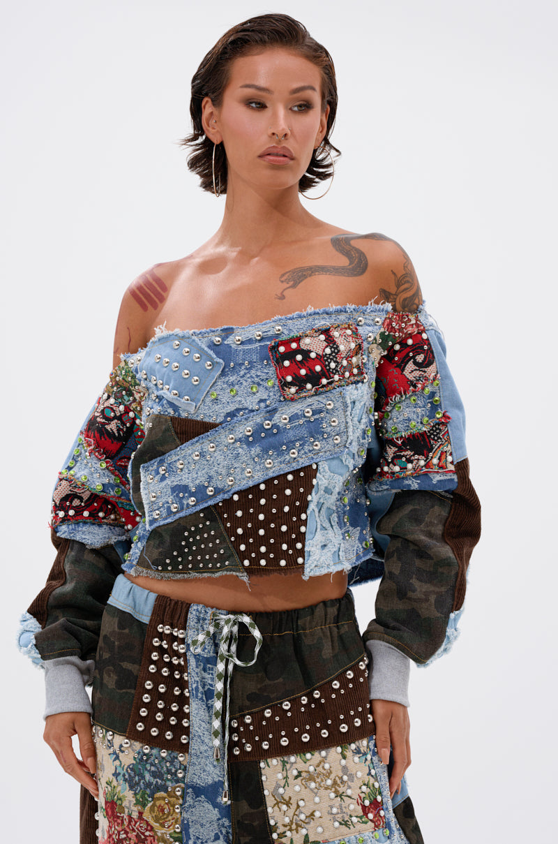 A woman with short, slicked-back hair models the CALL ME CRAZY PATCHWORK OFF THE SHOULDER SWEATSHIRT and matching studded pants—featuring denim, florals, corduroy, and camo—against a plain light background.