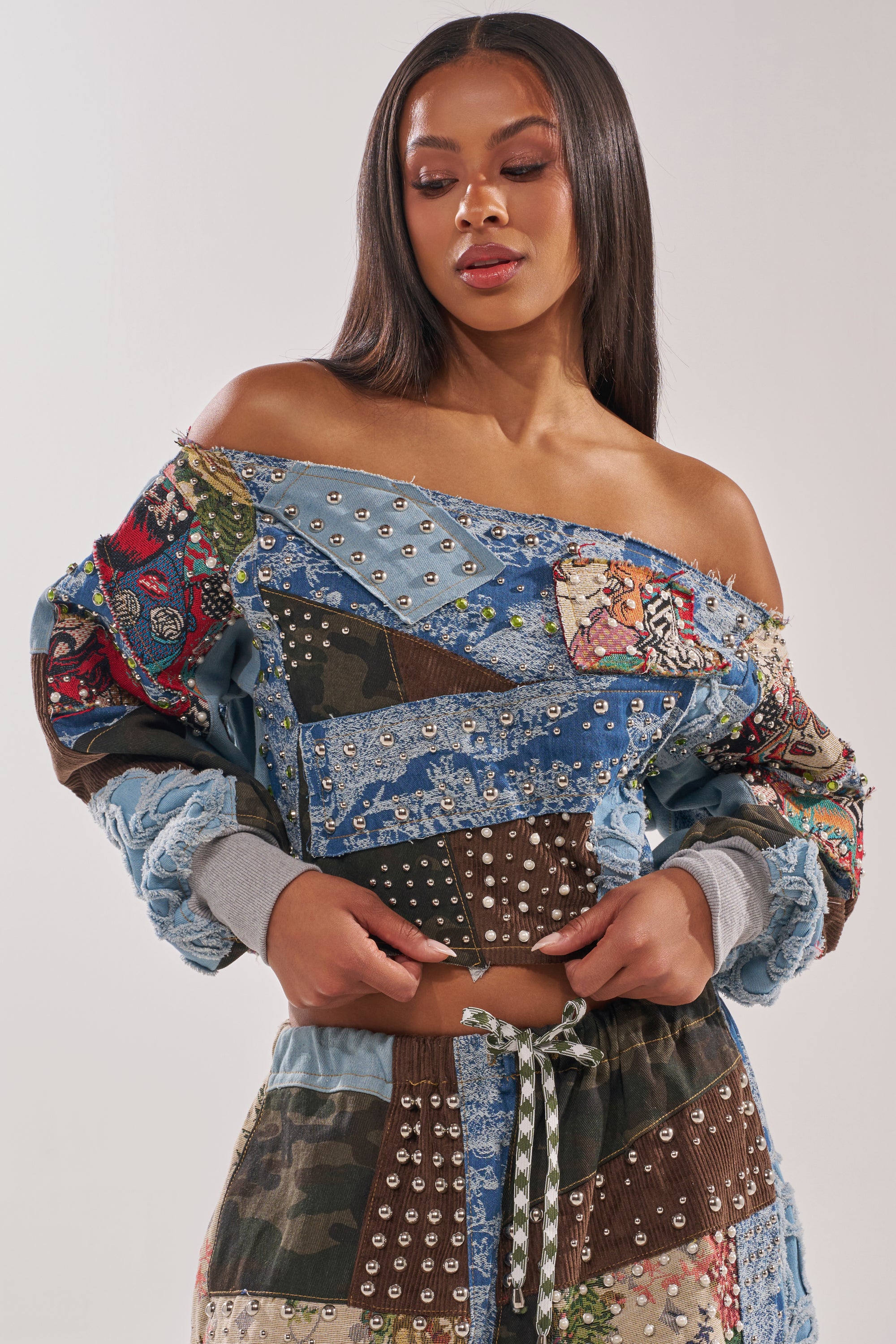 A woman in striking streetwear stands against a plain background, wearing the CALL ME CRAZY PATCHWORK OFF THE SHOULDER SWEATSHIRT and matching pants with mixed patterns, studs, and drawstrings. She looks down with her hands at her waist. 1