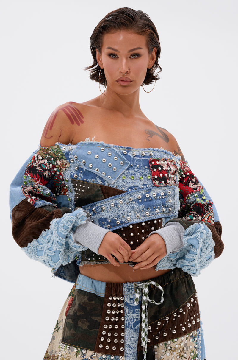 A person with short hair rocks an eclectic streetwear style, featuring the CALL ME CRAZY PATCHWORK OFF THE SHOULDER SWEATSHIRT with bold patterns. Large hoop earrings and visible tattoos complete the edgy look.