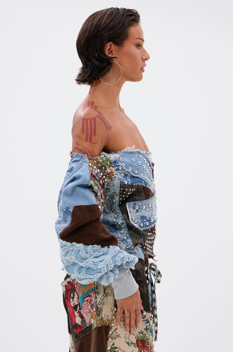 A woman with short dark hair stands in profile against a white background, wearing the CALL ME CRAZY PATCHWORK OFF THE SHOULDER SWEATSHIRT with denim, lace, pearl and sequin details. She has a tattoo and hoop earrings.