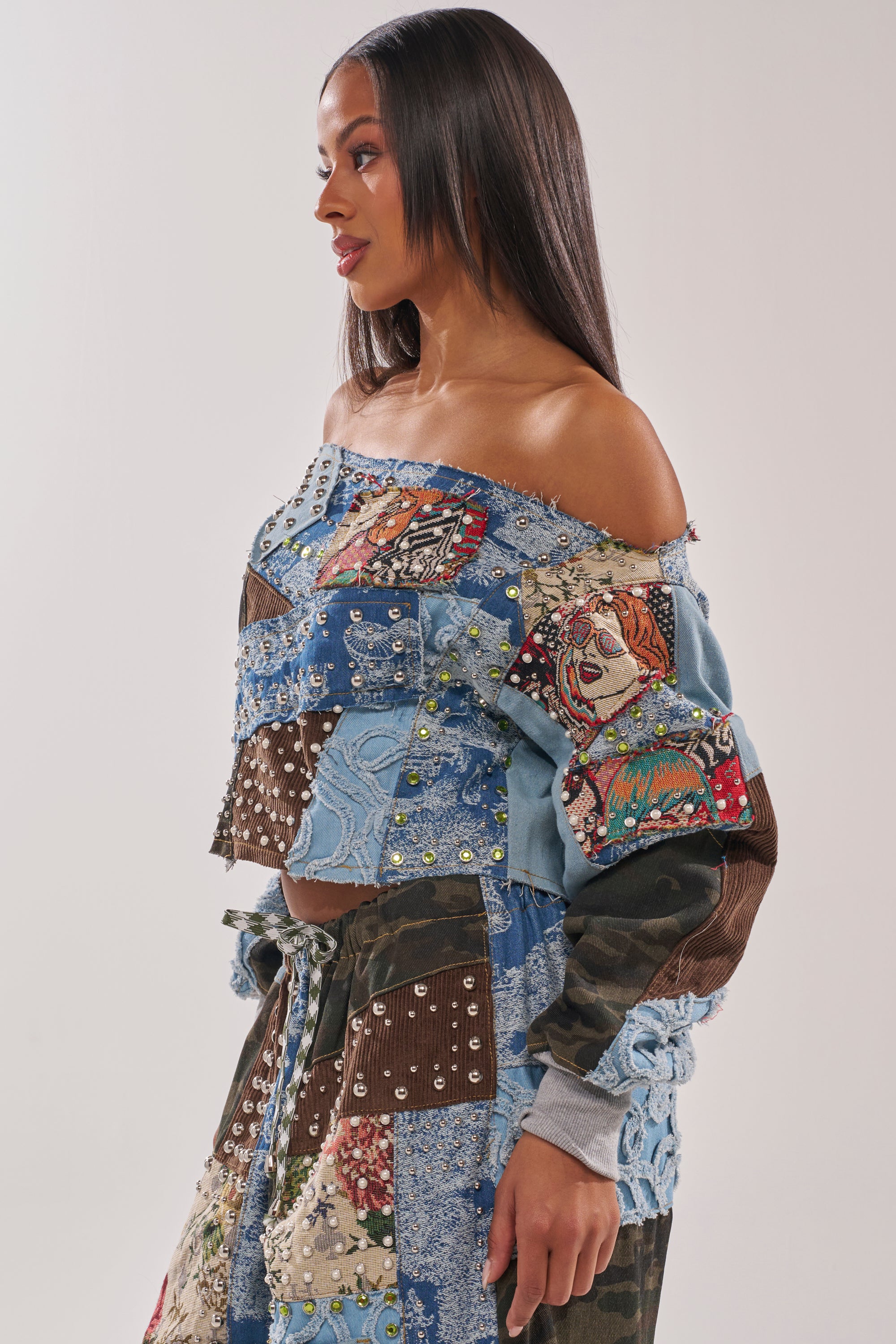 A woman with long dark hair models the CALL ME CRAZY PATCHWORK OFF THE SHOULDER SWEATSHIRT, featuring colorful fabric, beaded details, and face prints. She stands sideways against a plain background, showcasing unique streetwear style. 3