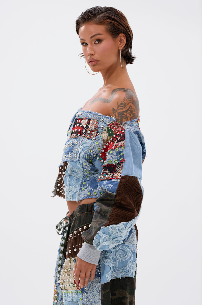 A woman with short hair and hoop earrings poses against a white backdrop, wearing the CALL ME CRAZY PATCHWORK OFF THE SHOULDER SWEATSHIRT and pants, her shoulder tattoo visible for an edgy streetwear vibe.