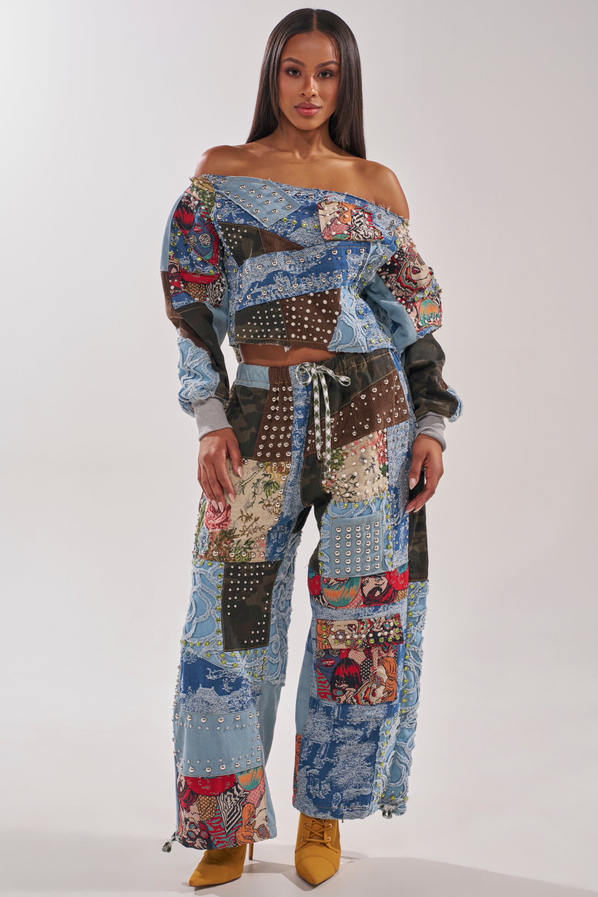 A woman wears the CALL ME CRAZY PATCHWORK OFF THE SHOULDER SWEATSHIRT paired with matching wide-leg pants, both featuring colorful patchwork denim and studded details. She finishes her vibrant streetwear look with mustard yellow heeled shoes. 6