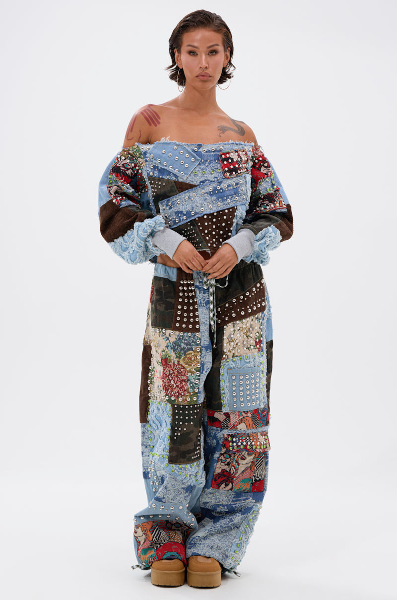 A person stands against a white background in eclectic streetwear, wearing the CALL ME CRAZY PATCHWORK OFF THE SHOULDER SWEATSHIRT with blue, red, and floral panels, silver studs, platform shoes, and wet slicked-back hair.