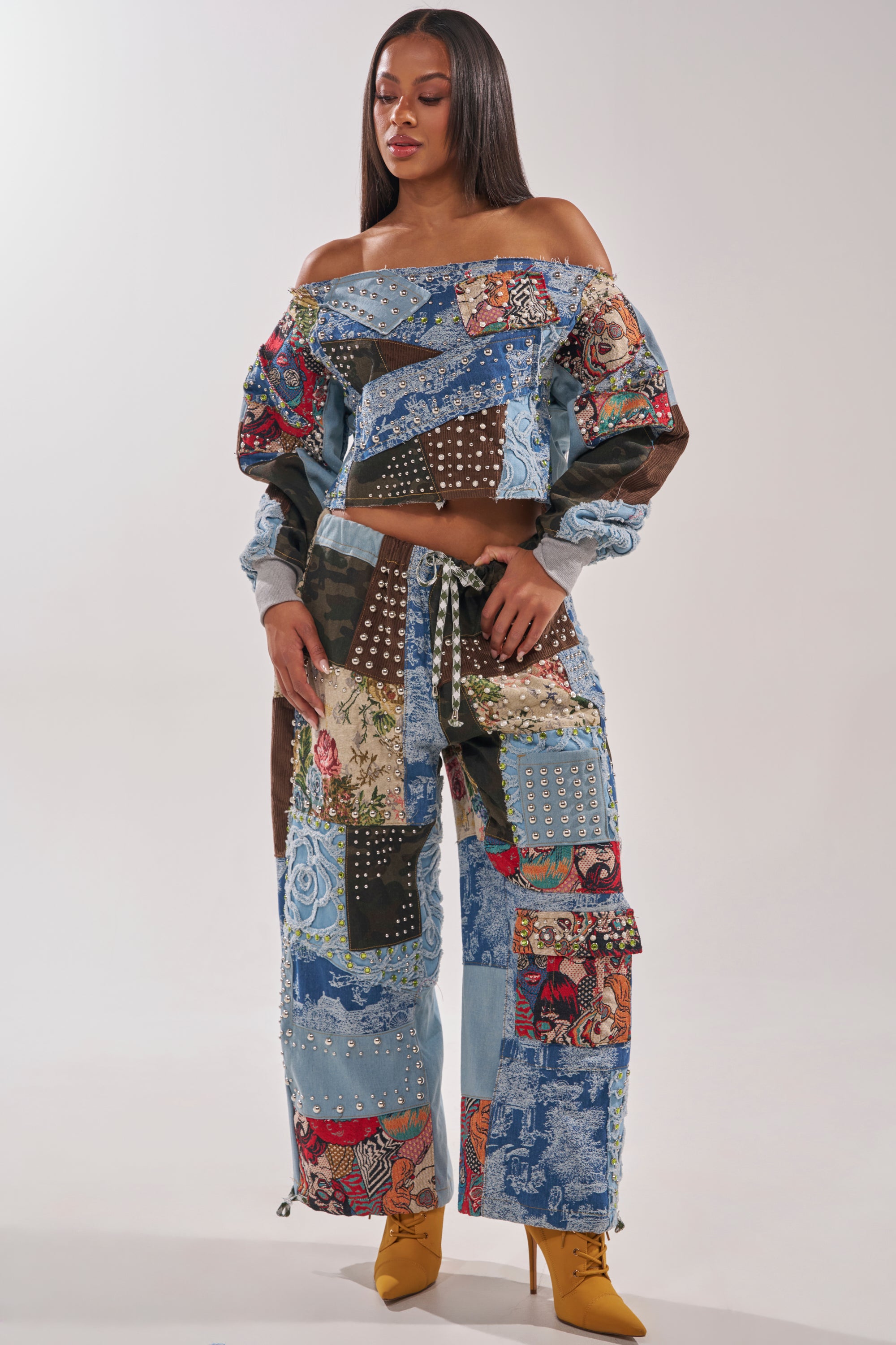 A woman wears the CALL ME CRAZY PATCHWORK OFF THE SHOULDER SWEATSHIRT and studded wide-leg pants, styled with straight hair and yellow heeled boots, set against a plain background. 7