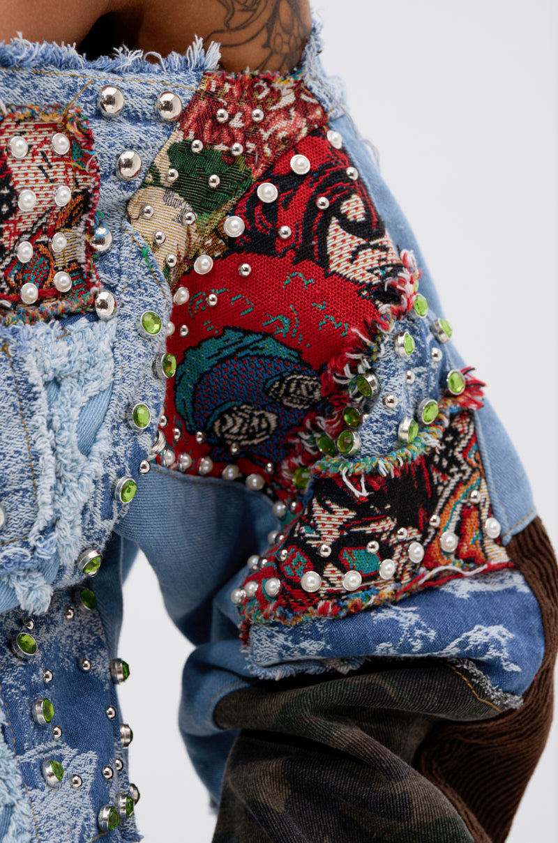 A close-up of a person in the CALL ME CRAZY PATCHWORK OFF THE SHOULDER SWEATSHIRT, featuring eclectic denim patches, colorful tapestry fabric, green and silver studs, pearls, and a visible shoulder tattoo that enhances its unique vibe.