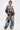 Wearing the CALL EM CRAZY PATCHWORK JOGGER with colorful patterns, a person with short brown hair pairs it with a studded patchwork denim off-the-shoulder top and tan shoes, posing confidently against a plain white background.