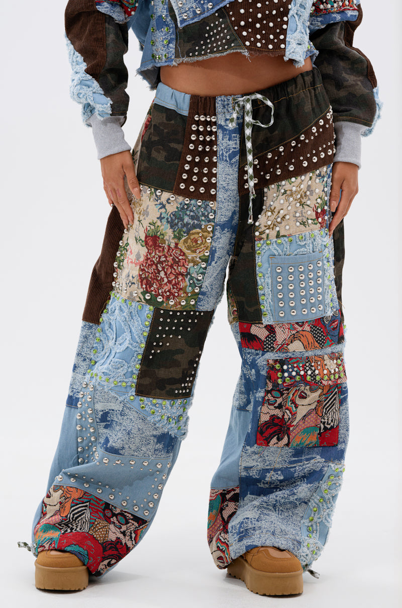 A person wears the CALL EM CRAZY PATCHWORK JOGGER—streetwear-inspired, high-rise joggers featuring a drawstring waist and wide-leg silhouette with denim, camo, floral, lace, and studded patches—paired with tan platform shoes.