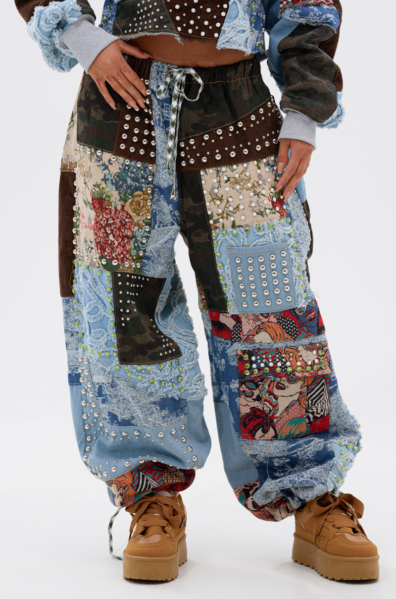 Someone stands against a plain white background wearing the CALL EM CRAZY PATCHWORK JOGGER featuring mixed fabric patterns, denim, and silver studs, paired with matching patchwork outerwear and tan platform shoes.