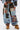 Someone stands against a plain white background wearing the CALL EM CRAZY PATCHWORK JOGGER featuring mixed fabric patterns, denim, and silver studs, paired with matching patchwork outerwear and tan platform shoes.