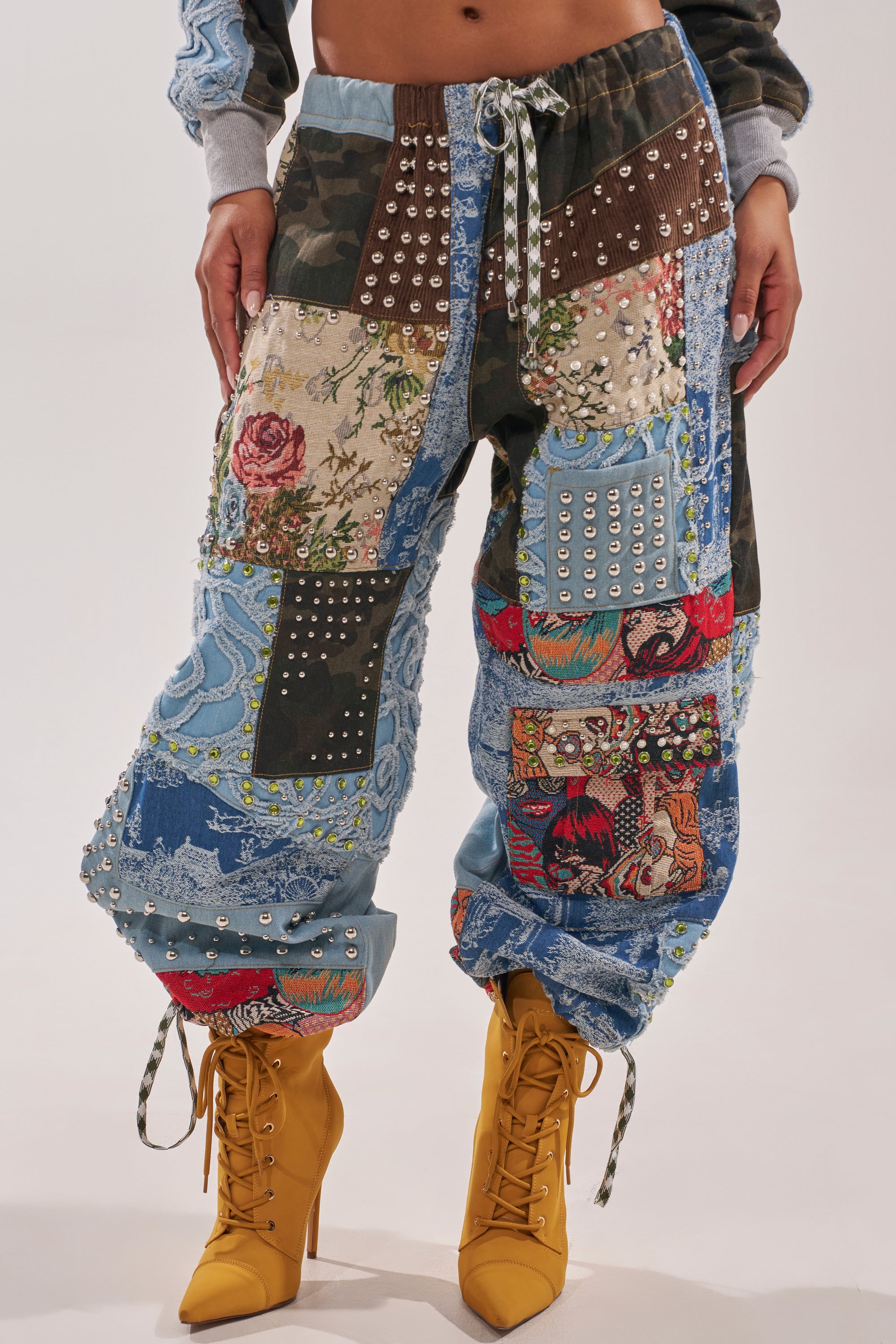 Wearing the CALL EM CRAZY PATCHWORK JOGGER with vibrant fabrics, patterns, studs, and embroidery, paired with mustard-yellow lace-up heeled boots. Only the lower half shows—ideal for festival jogger style. 2