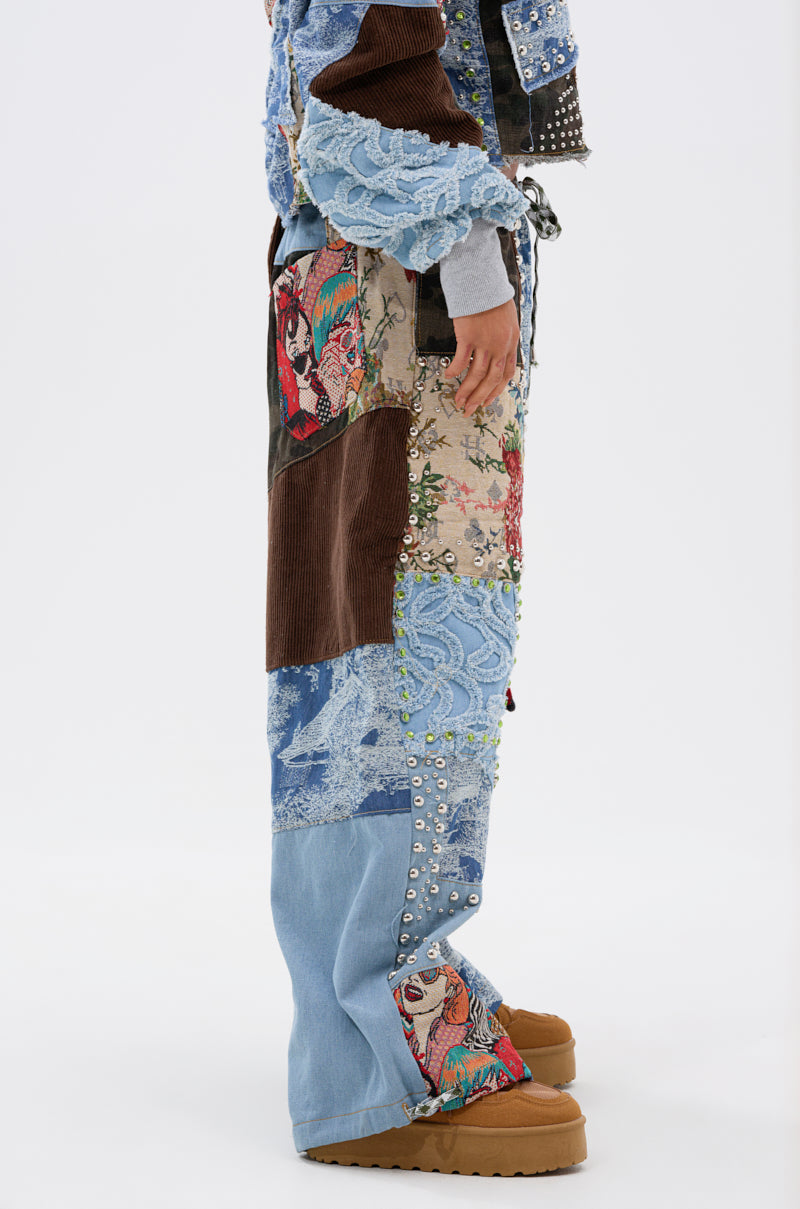 A person stands in profile against a plain white background wearing the CALL EM CRAZY PATCHWORK JOGGER, featuring denim, lace, corduroy, floral prints with metal studs and embroidery, paired with tan platform shoes.