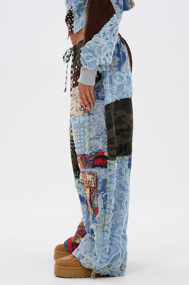 Side view of a person wearing CALL EM CRAZY PATCHWORK JOGGER, showcasing wide leg joggers in textured denim, lace, camouflage, and patterned patches with tan platform shoes for a bold streetwear look against a white background.