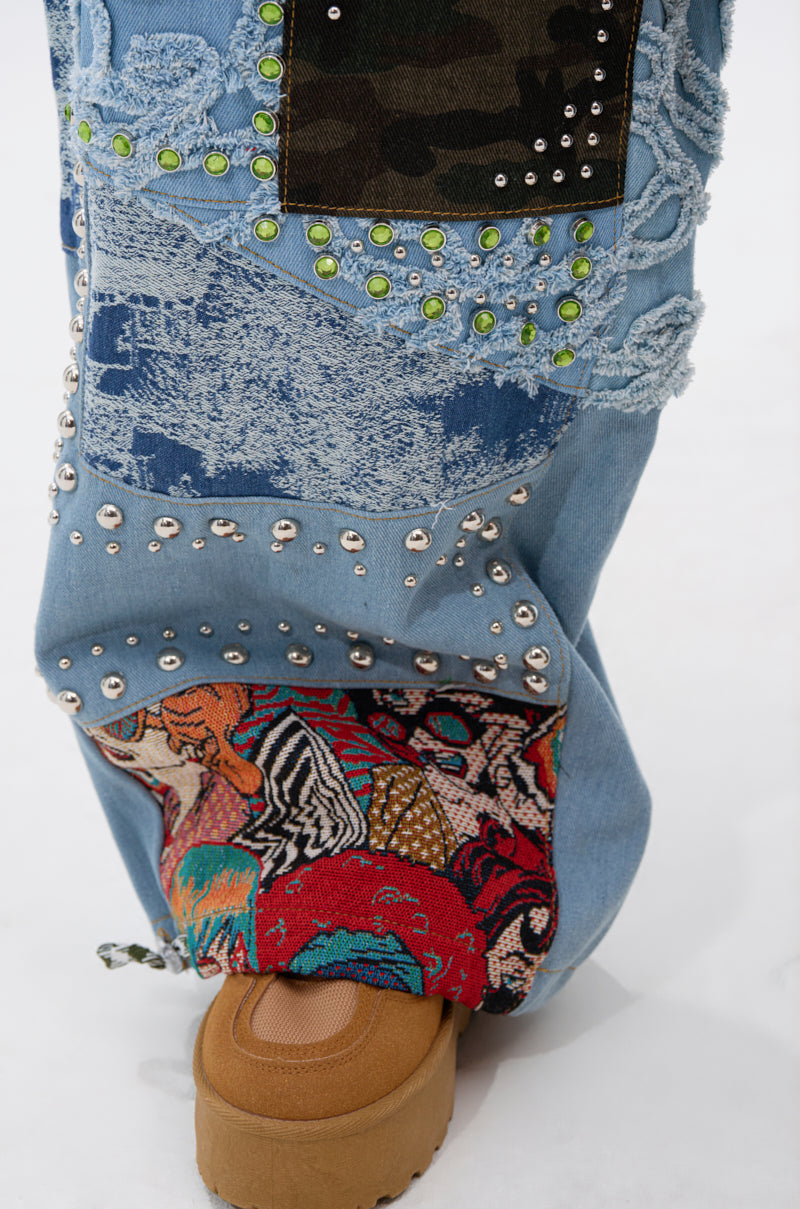 A close-up of the CALL EM CRAZY PATCHWORK JOGGER showcases denim, camo, and colorful patches with silver studs and green beads, styled with a tan platform shoe against a white background.