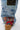 A close-up of the CALL EM CRAZY PATCHWORK JOGGER showcases denim, camo, and colorful patches with silver studs and green beads, styled with a tan platform shoe against a white background.