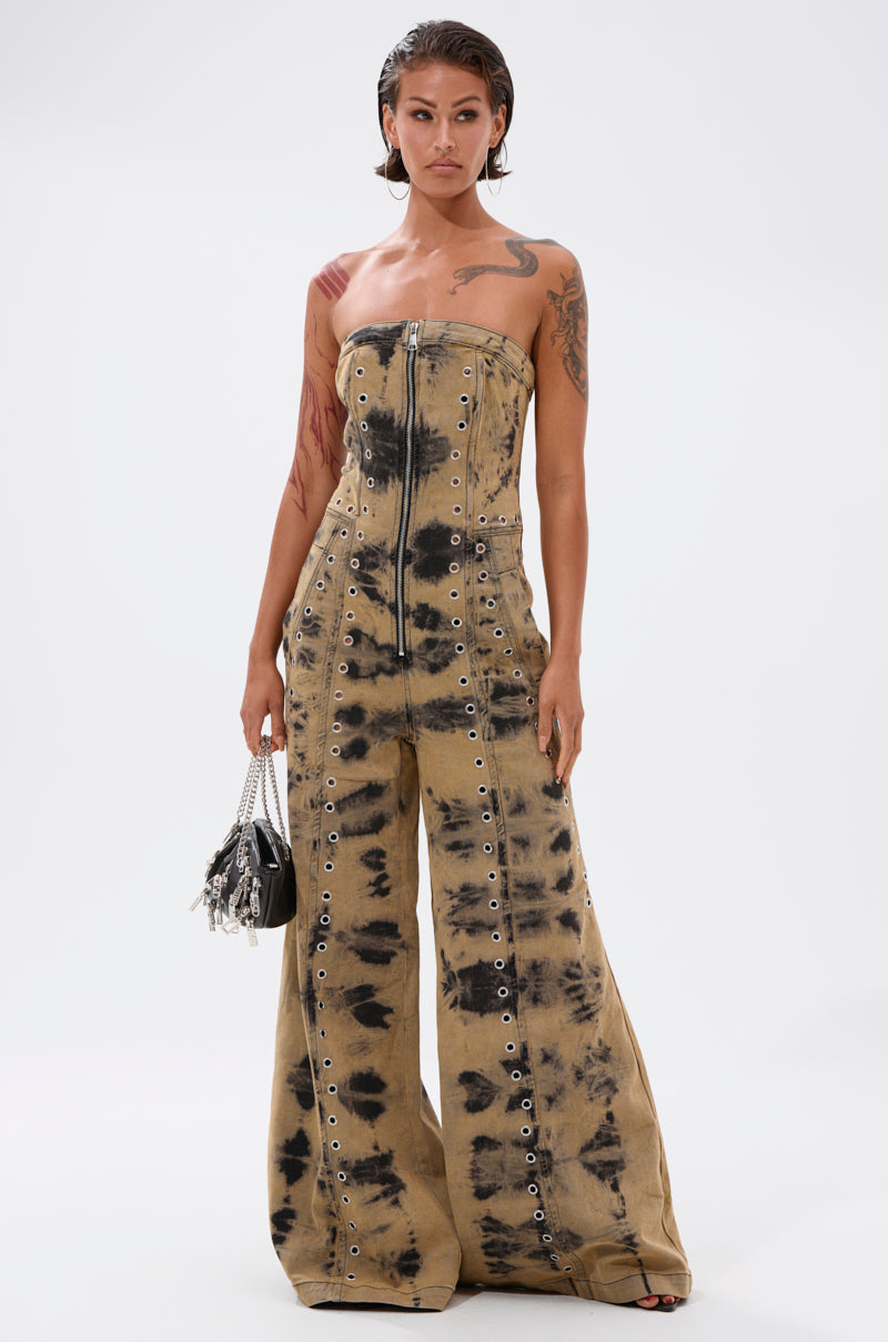 A person with wet, slicked-back hair wears the INTERNET FAMOUS PAINTED DENIM JUMPSUIT, featuring a strapless design with front zipper and grommet details, holding a small black handbag against a plain white background.