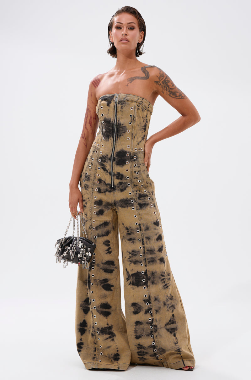 A person with short hair and tattoos wears the INTERNET FAMOUS PAINTED DENIM JUMPSUIT with metal eyelet details, holding a small studded handbag against a plain white background.