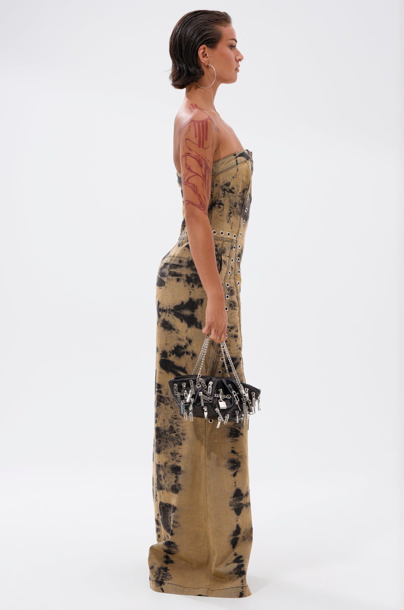 A woman stands in profile wearing the INTERNET FAMOUS PAINTED DENIM JUMPSUIT, a strapless design with metal eyelets. She holds a black handbag with silver charms, has slicked-back hair, and sports a large upper arm and shoulder tattoo.