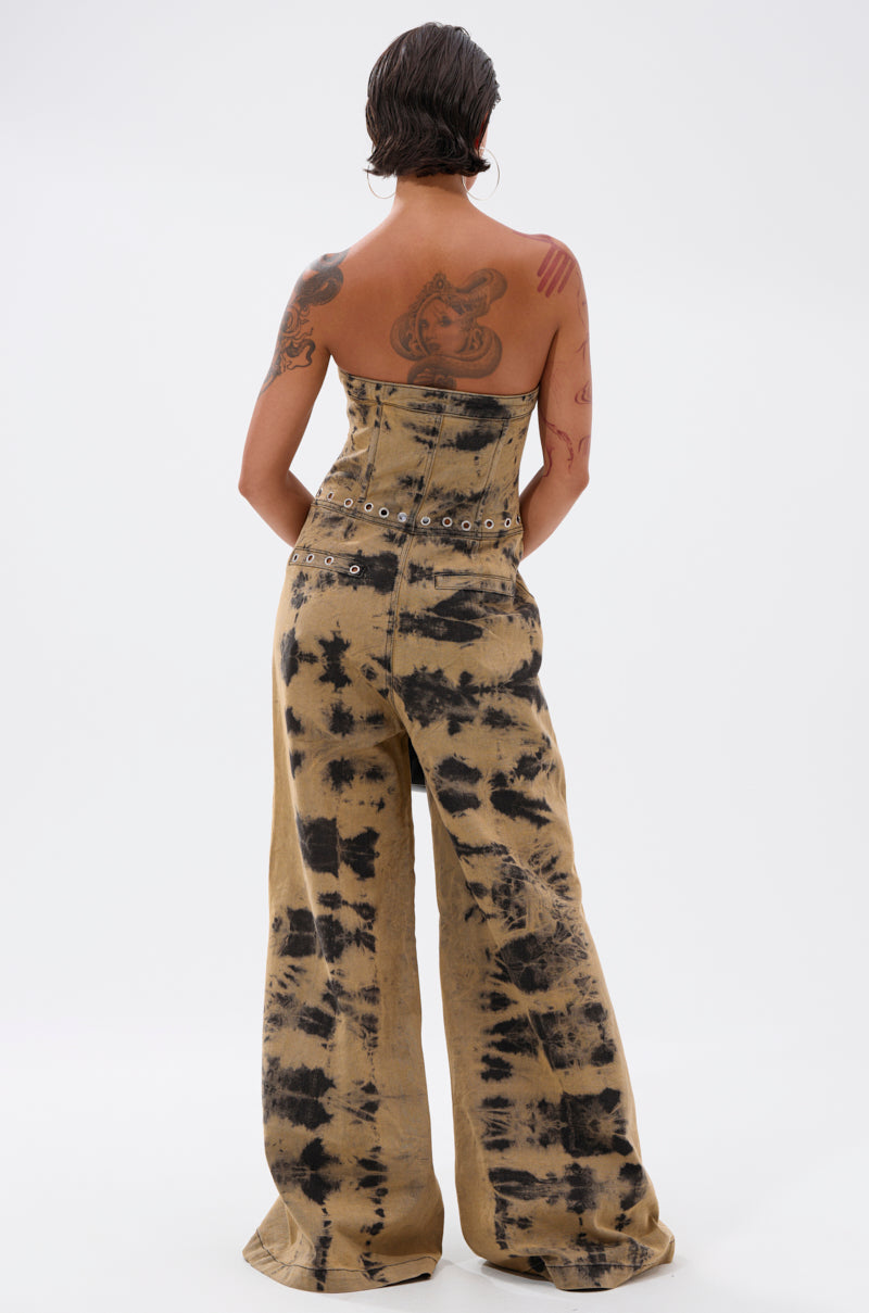 A person with short hair and visible tattoos stands with their back to the camera, wearing the strapless INTERNET FAMOUS PAINTED DENIM JUMPSUIT in tan and black, featuring wide legs and a belt with circular metal eyelets.