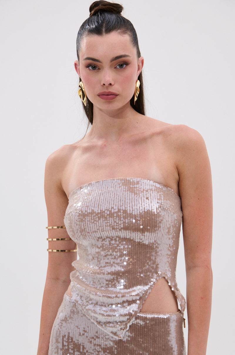 A woman wears the SPARKLE QUEEN SEQUIN BLOUSE with shimmering sequins and a metallic back zipper. Her long dark hair is in a high bun, accessorized with gold hoop earrings and an arm cuff, against a plain light background.