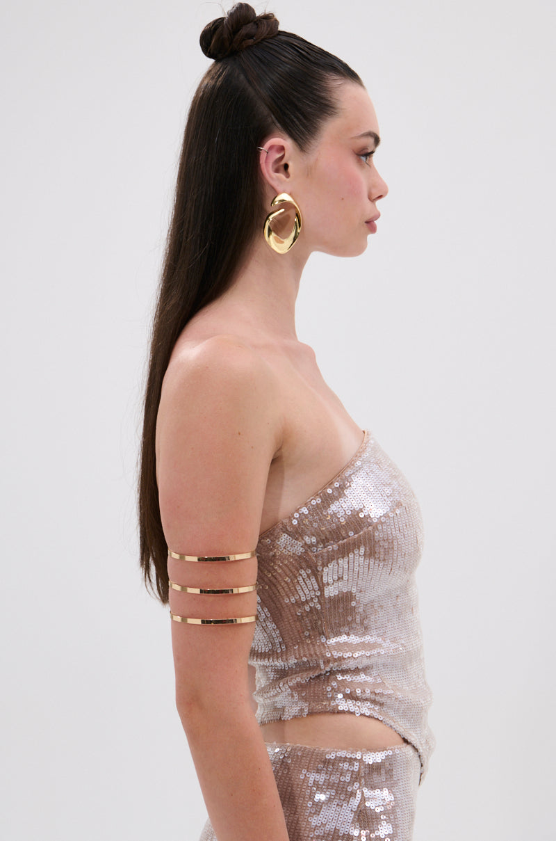 A woman with long, straight hair in a topknot bun wears the SPARKLE QUEEN SEQUIN BLOUSE with a metallic back zipper, accessorized with large gold hoops and a gold armband, posing in profile against a plain background.