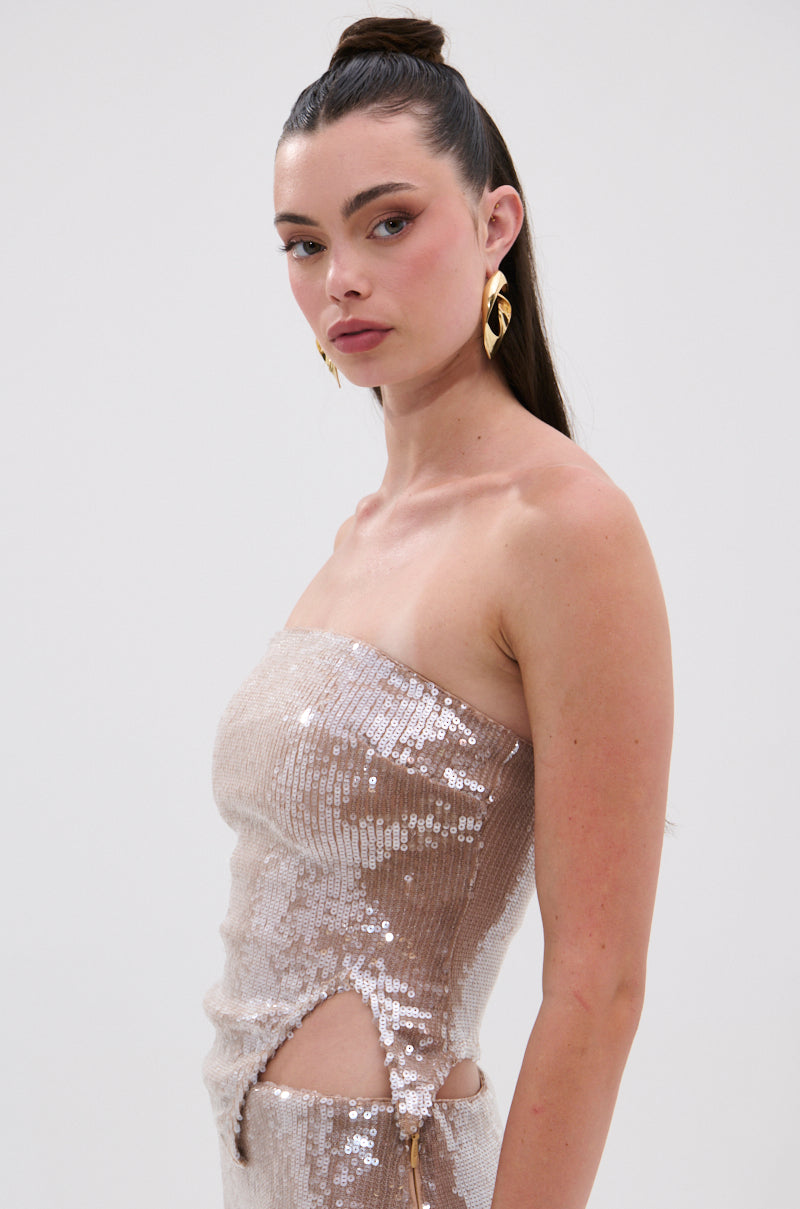 A woman with long brown hair in a high ponytail wears gold earrings and the SPARKLE QUEEN SEQUIN BLOUSE, featuring a strapless, sequined beige design with side cutouts and a metallic back zipper. She looks confidently at the camera against a plain background.