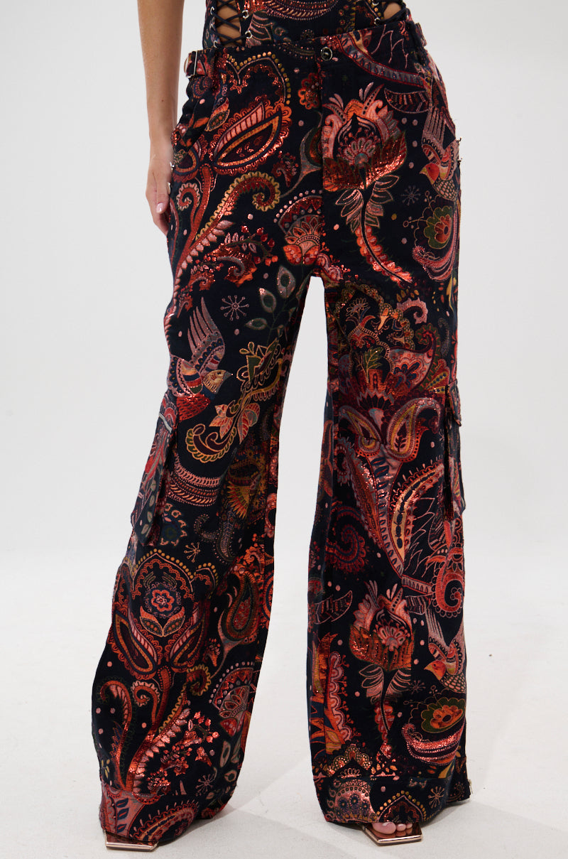 A person models the FALL VIBES BROCADE TROUSER—ultra-wide leg pants featuring a vibrant red, orange, and pink paisley pattern on black—cropped from the waist down against a plain white background.