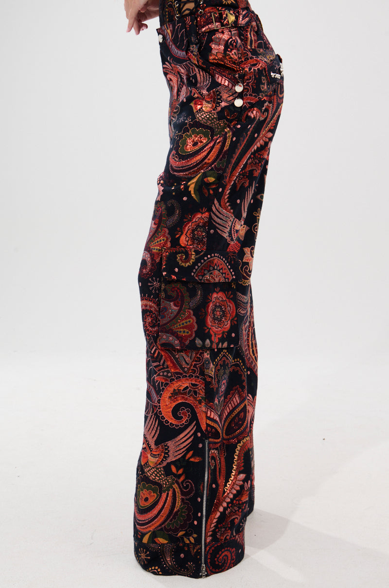 A person stands sideways modeling the FALL VIBES BROCADE TROUSER, which features an ultra-wide leg silhouette and a vibrant paisley and floral pattern with metallic red, orange, and black tones against a white background.