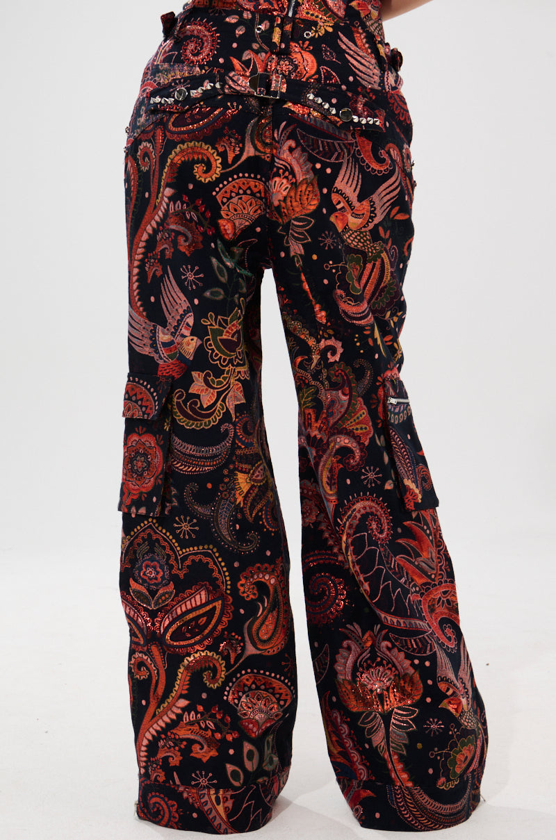 A person wears the FALL VIBES BROCADE TROUSER—high-rise, wide-leg pants with a vibrant multicolored paisley and floral print. Only their lower half is visible against a plain white background.