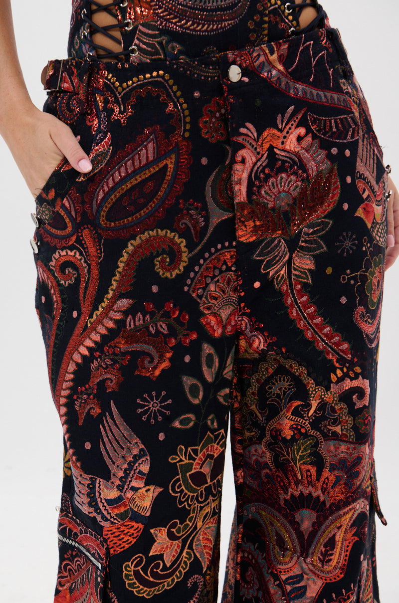 A person models the FALL VIBES BROCADE TROUSER, showcasing its bold, metallic autumnal print in red, orange, and gold. Their hand rests in the right pocket of the ultra-wide leg silhouette.