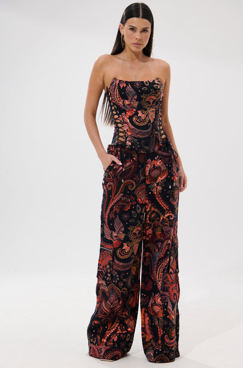 A woman stands against a white background wearing the FALL VIBES BROCADE CORSET WITH LACE UP SIDES, featuring an autumnal paisley print in red, orange, and black. She has long straight hair and her hands in her pockets.