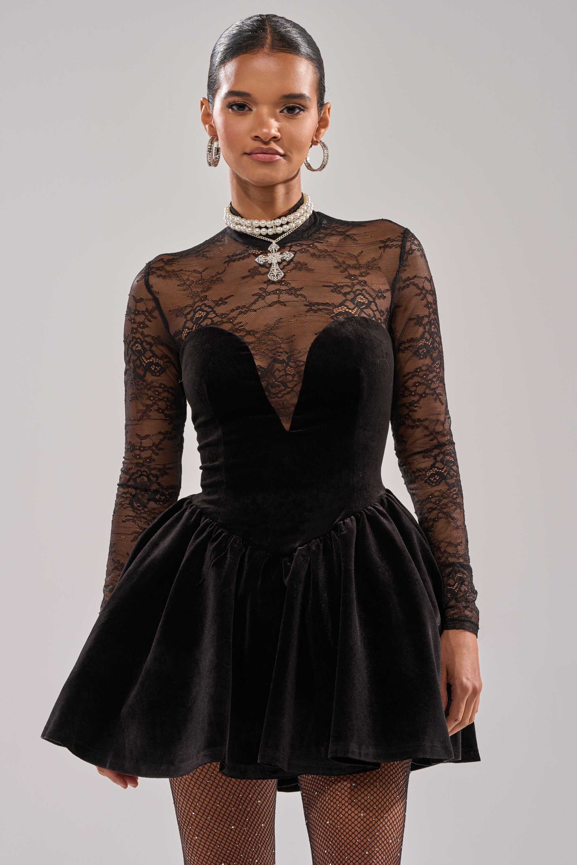A woman wears the DRESSED TO IMPRESS LACE MINI DRESS, featuring a velvet black fabric, sheer lace top, long sleeves, and flared skirt. She styles it with pulled-back hair, hoop earrings, a jeweled necklace, and fishnet tights.