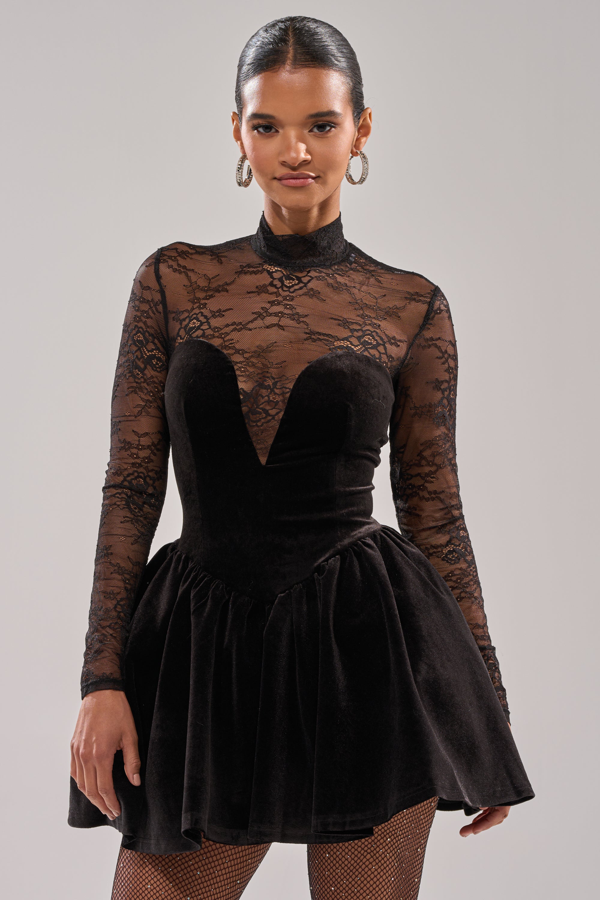 A woman stands facing forward in the DRESSED TO IMPRESS LACE MINI DRESS, featuring a velvet finish, lace bodice, long sleeves, and flared skirt. She accessorizes with hoop earrings and wears her hair slicked back against a plain background.