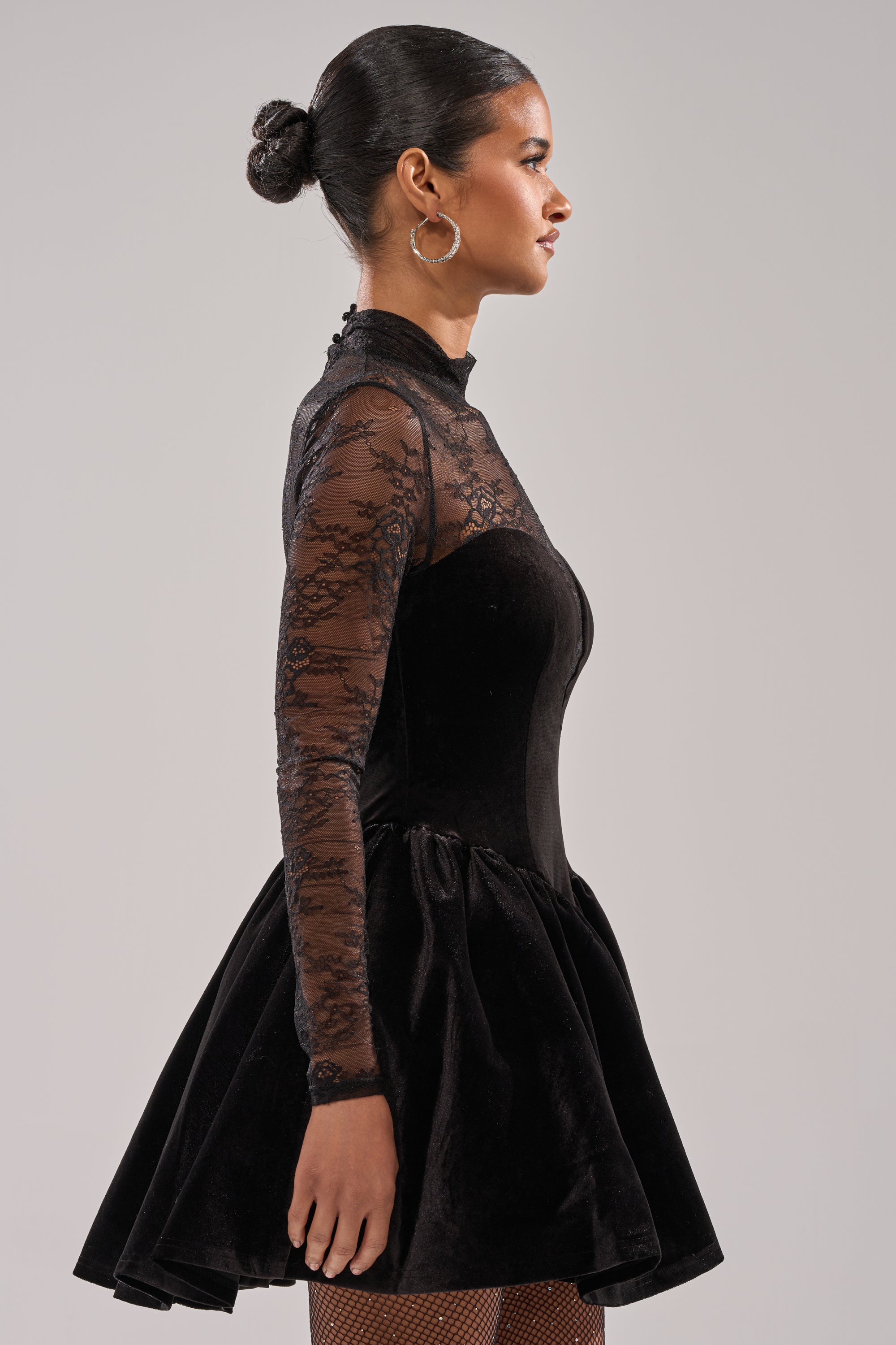A woman in profile wears the DRESSED TO IMPRESS LACE MINI DRESS in black velvet, featuring a floral lace bodice, sheer lace sleeves, and a flared skirt. Her hair is in a sleek bun with hoop earrings against a plain light-colored background.