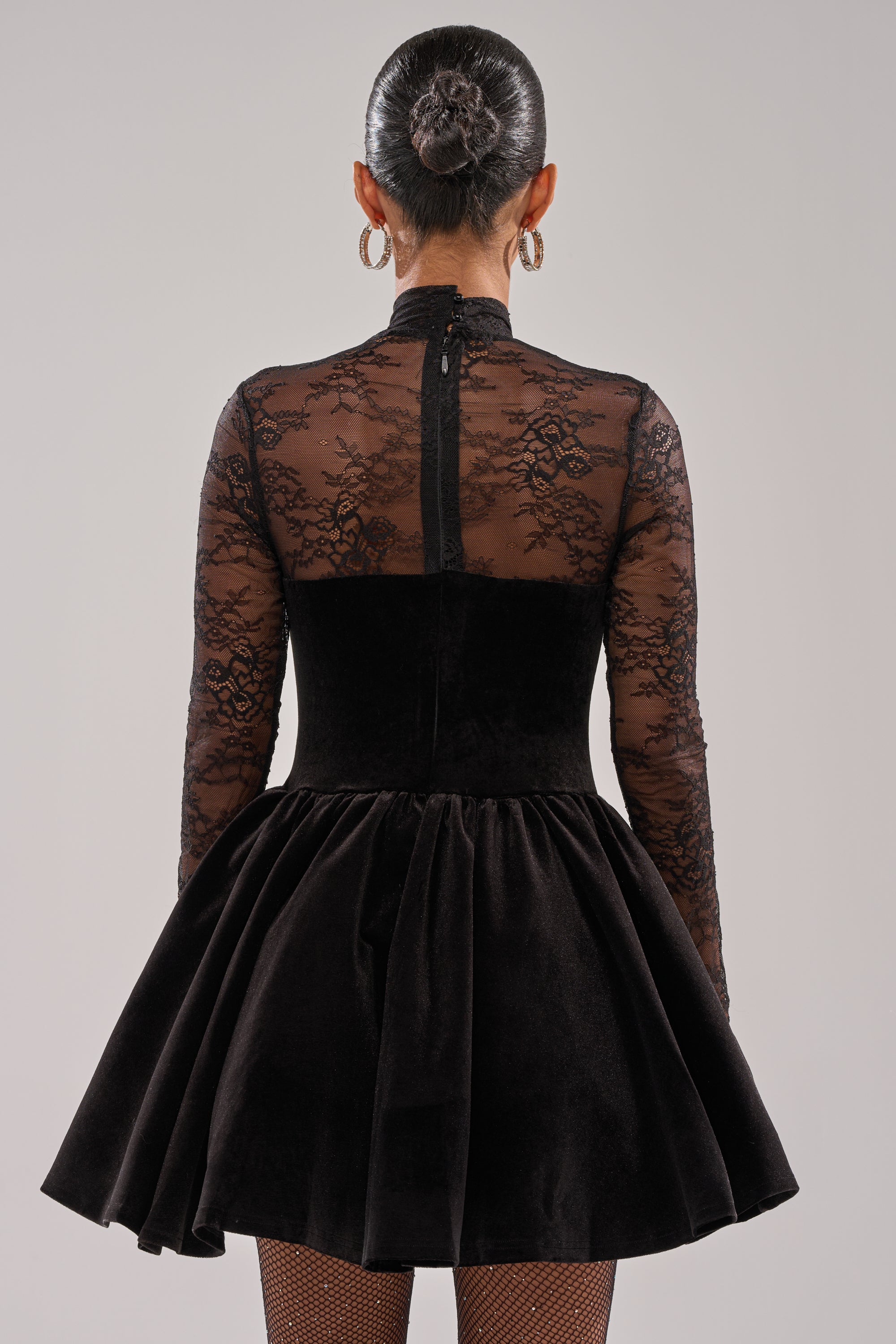 A person with their hair in a sleek bun wears the DRESSED TO IMPRESS LACE MINI DRESS, featuring a lace long-sleeve top and flared skirt. The back view showcases its elegant design against a plain light background.