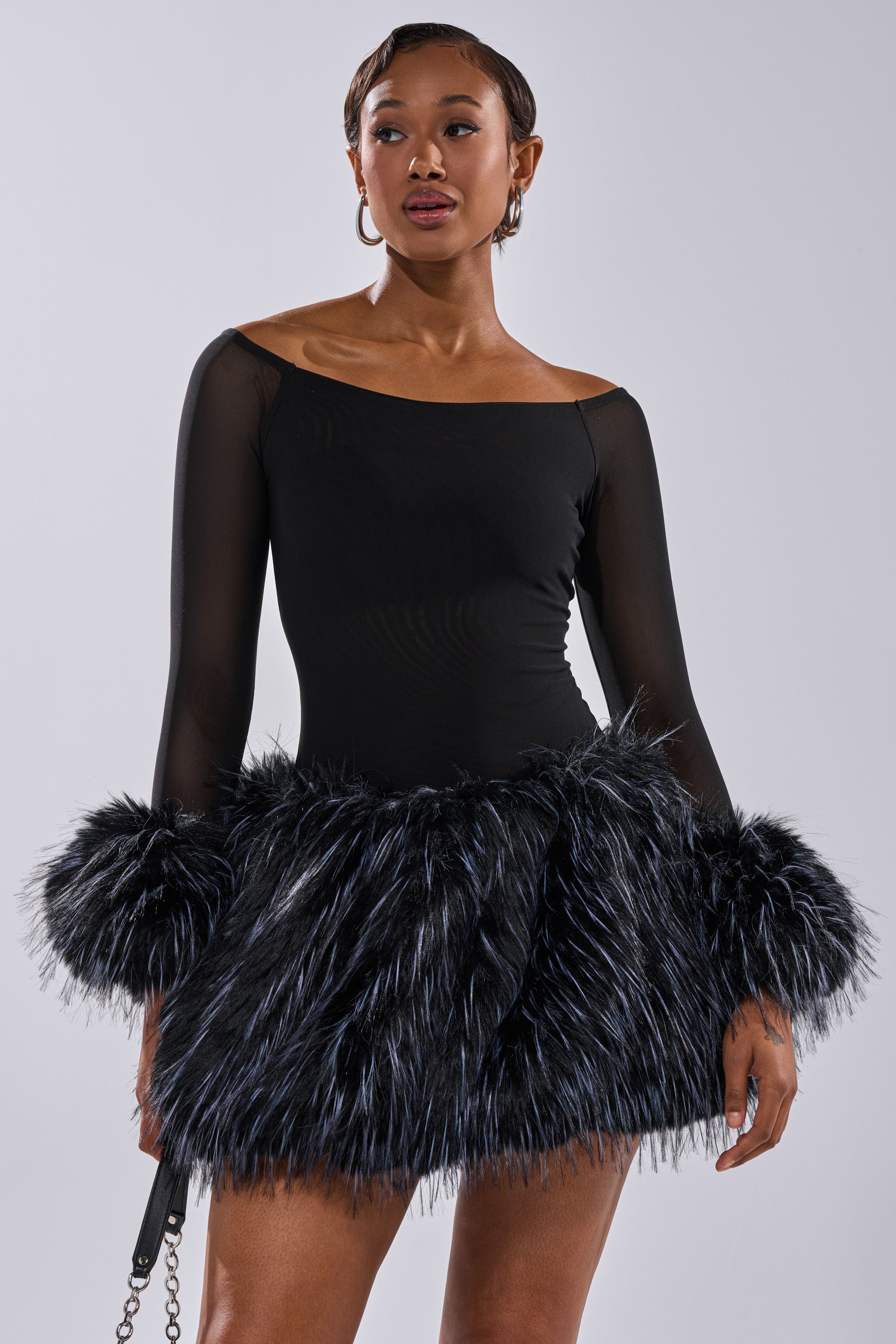 A woman wears the KELYN CALLED FUR MINI DRESS—an off-the-shoulder sheer mesh party dress with long sleeves and black and silver faux fur skirt and cuffs—posing against a plain light background.