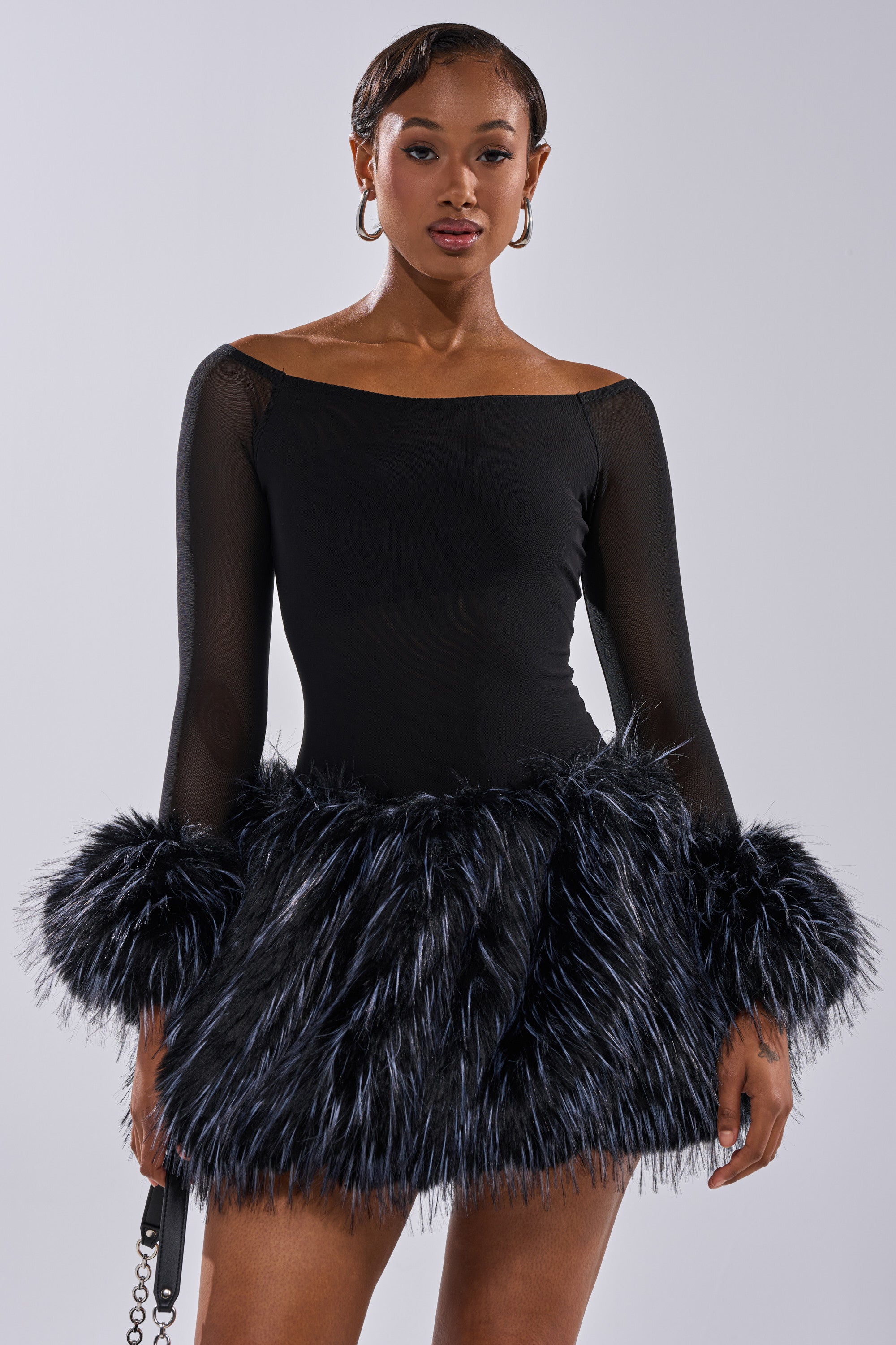 A woman poses confidently against a plain light background, wearing the KELYN CALLED FUR MINI DRESS—a stylish black party dress with sheer long sleeves and bold furry accents at the cuffs and skirt hem.