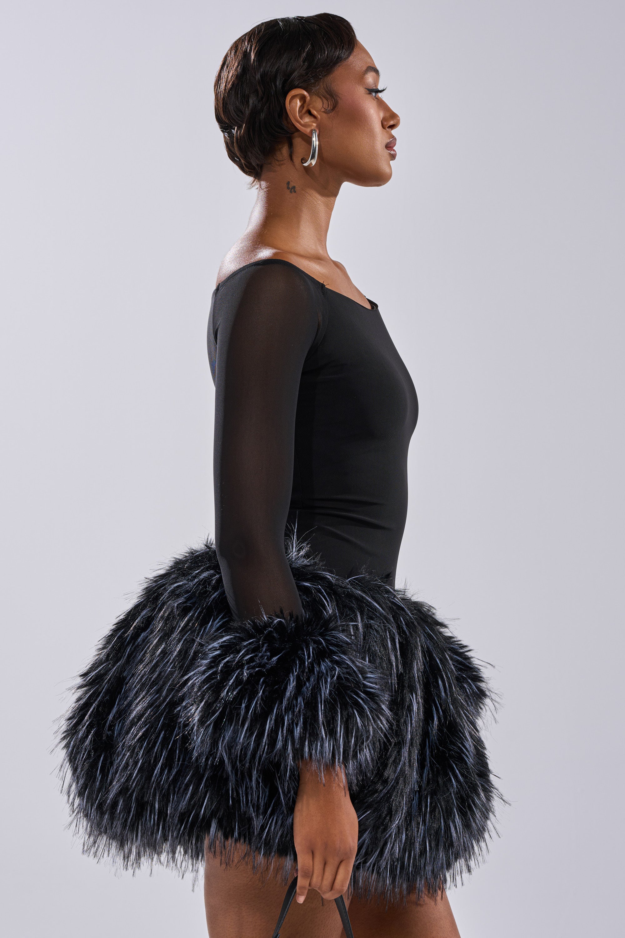A woman with short hair and large hoop earrings stands in profile wearing the KELYN CALLED FUR MINI DRESS, featuring sheer sleeves and a voluminous black faux fur mini skirt, holding a black strap in her hand.