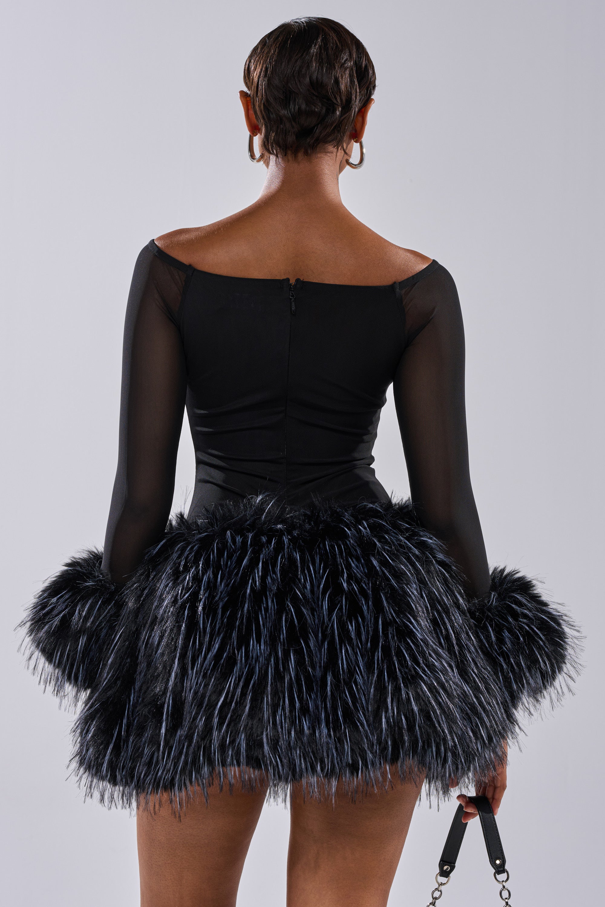 A person with short hair is shown from the back in the KELYN CALLED FUR MINI DRESS, a fitted black party dress with sheer long sleeves and a dramatic, fluffy feathered skirt, set against a plain gray background.
