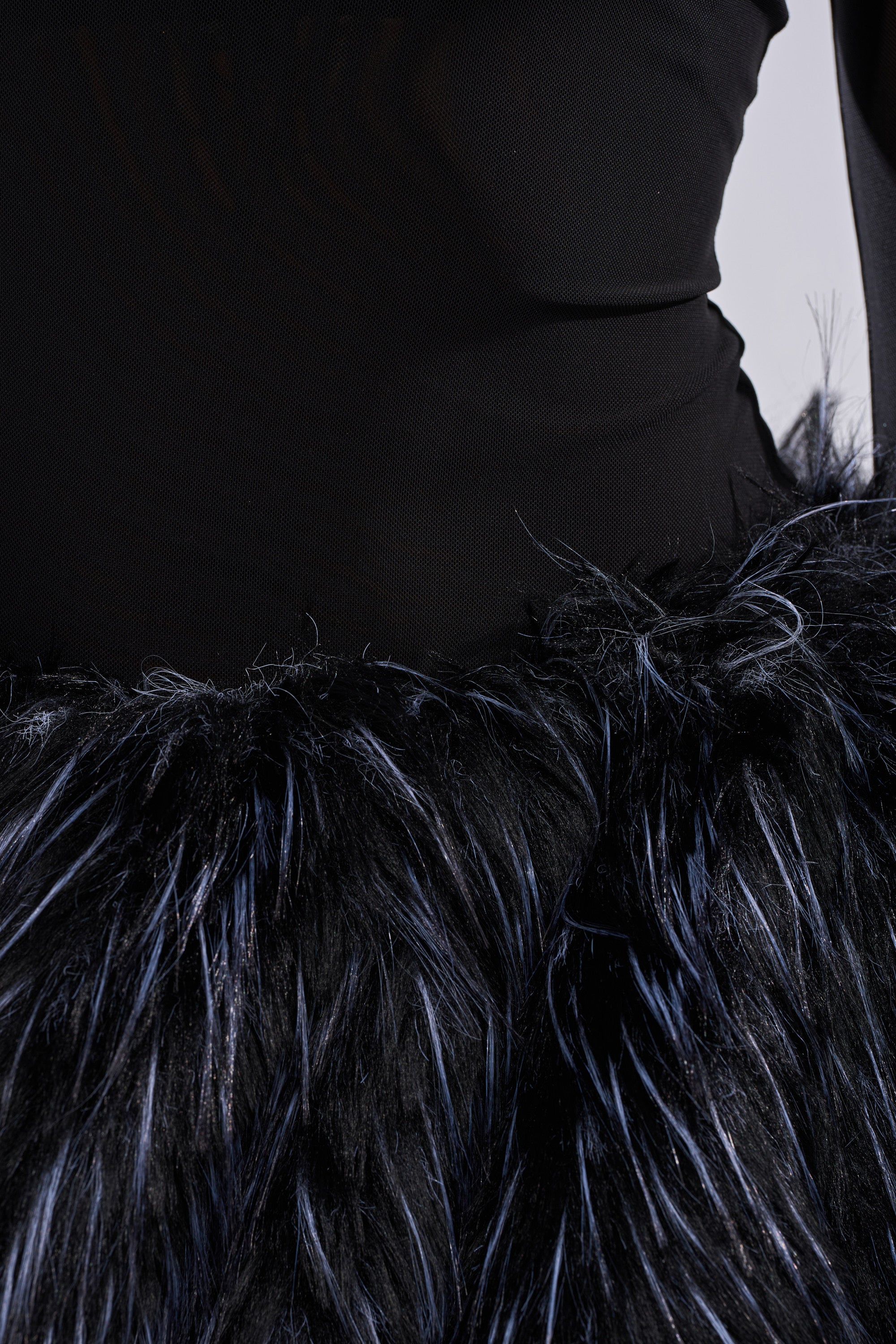 A close-up of a person wearing the KELYN CALLED FUR MINI DRESS—a black, fluffy mini dress with shiny, textured fur-like material—styled over a fitted black long-sleeved top for a chic party look.