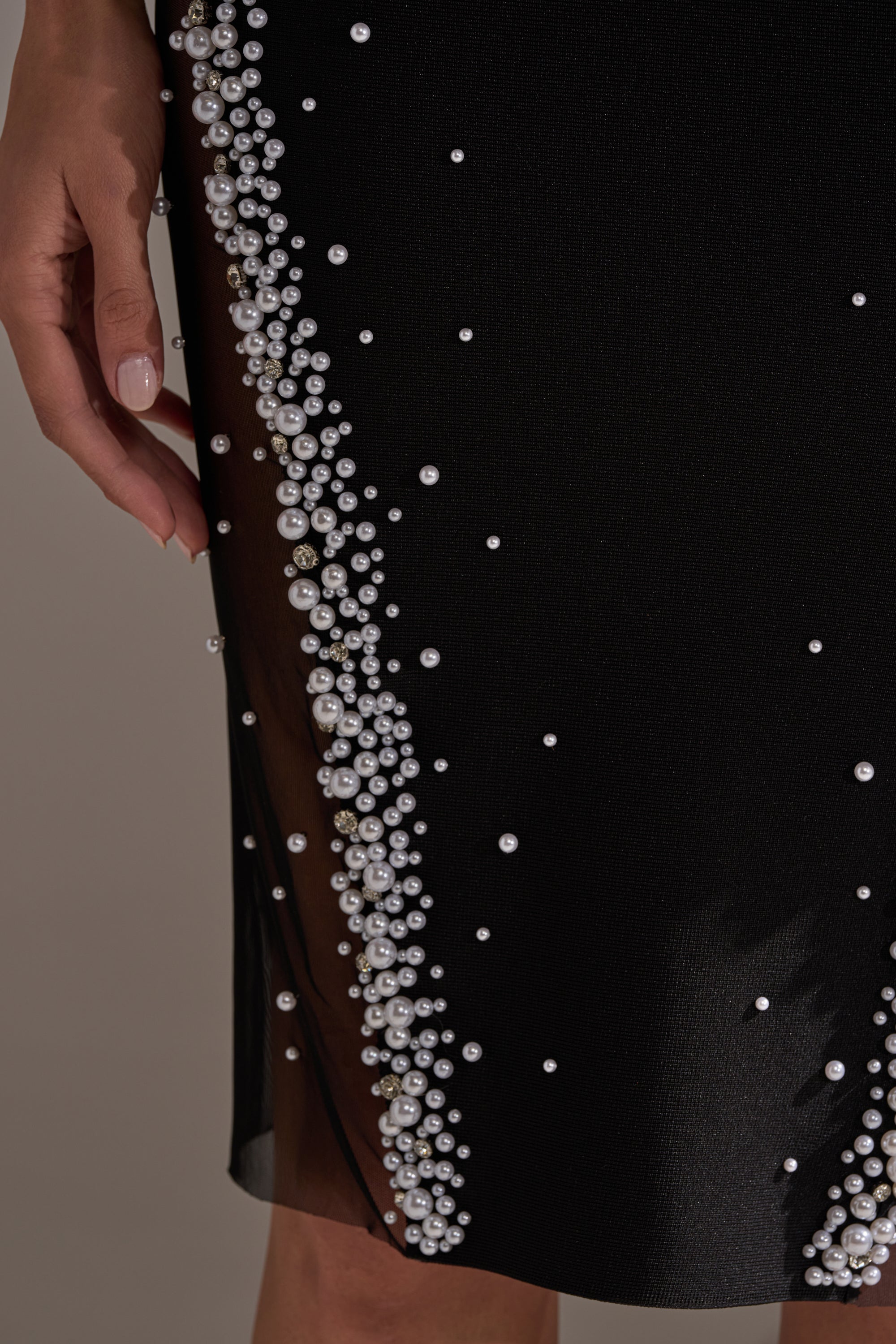 A close-up of a hand resting beside the CHAMPAGNE EDIT RHINESTONE MIDI DRESS, a black bandage fabric dress adorned with clusters of white pearls, small beads, and a sheer mesh panel.