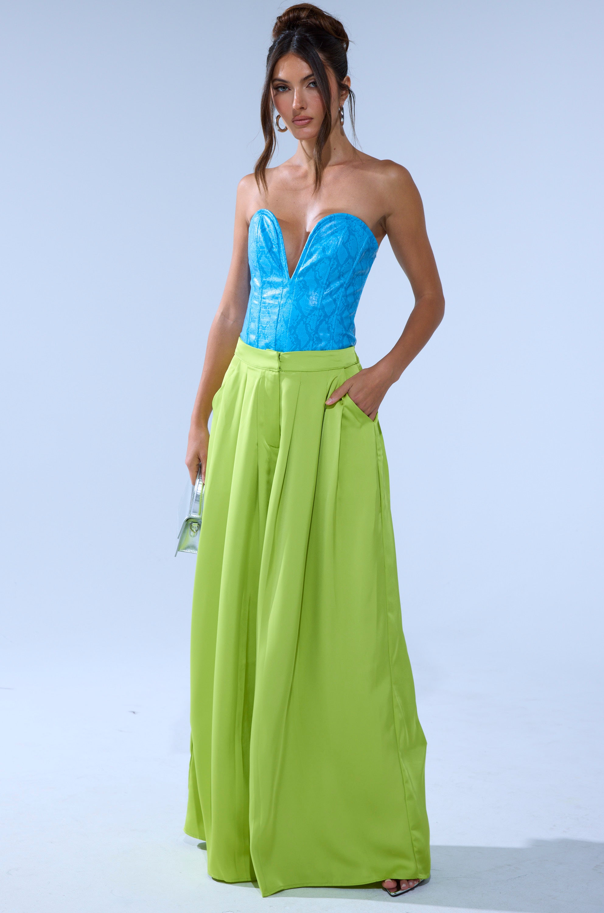A woman models the CHARTREUSE PLEATED PALAZZO PANT with a strapless blue corset top, clear purse, hoop earrings, and a loose updo against a plain light gray background. 0
