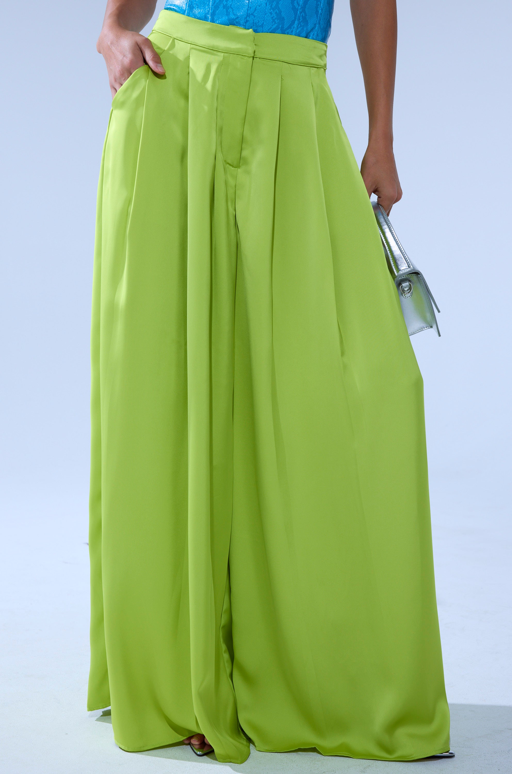 Wearing the CHARTREUSE PLEATED PALAZZO PANT with a wide-leg silhouette, a person pairs it with a blue top and carries a small silver handbag against a plain light background. 1