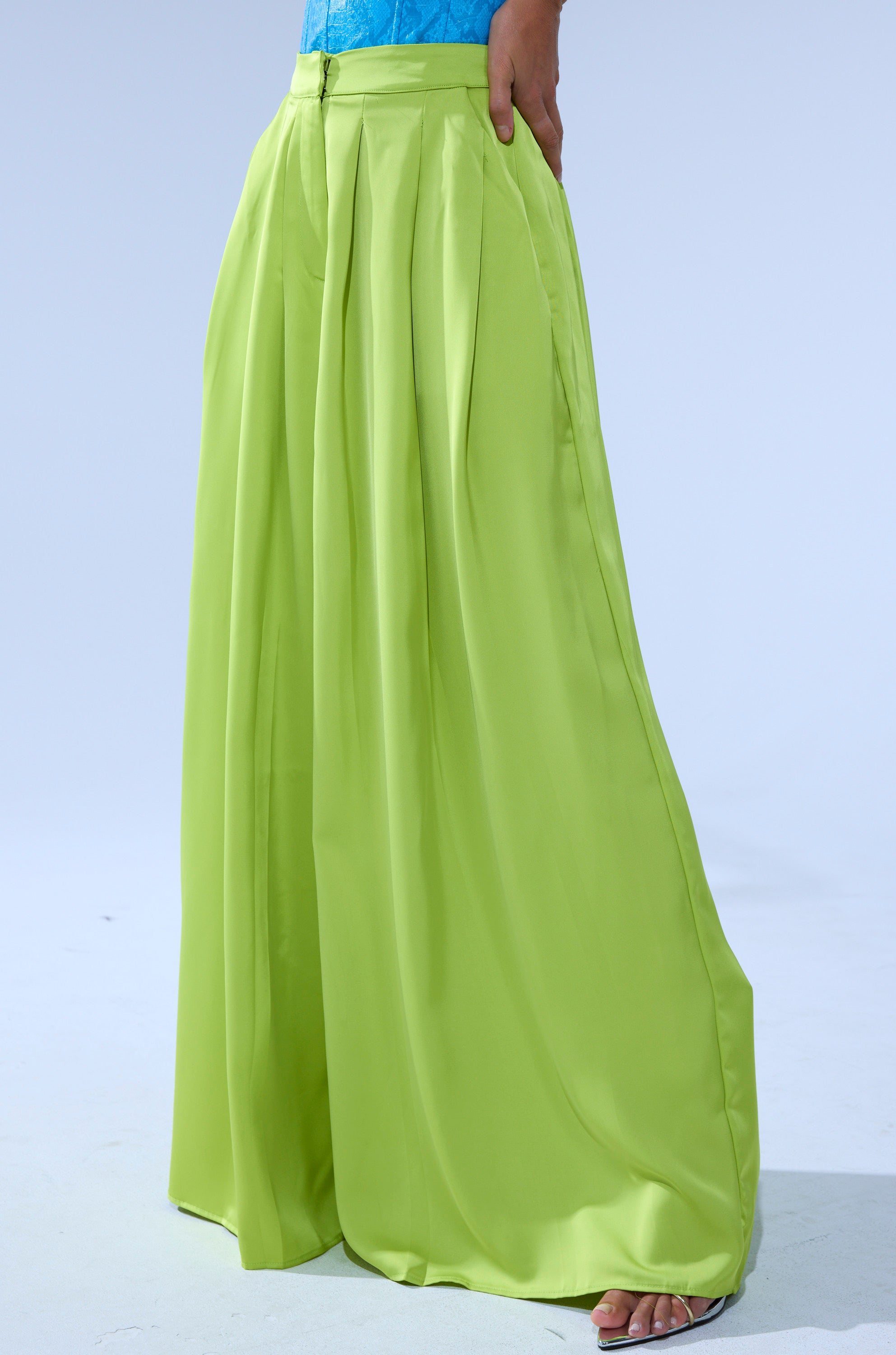 A person models the CHARTREUSE PLEATED PALAZZO PANT, featuring pleats and a wide-leg silhouette, paired with a turquoise top. Only their lower body is visible against a light background. 2