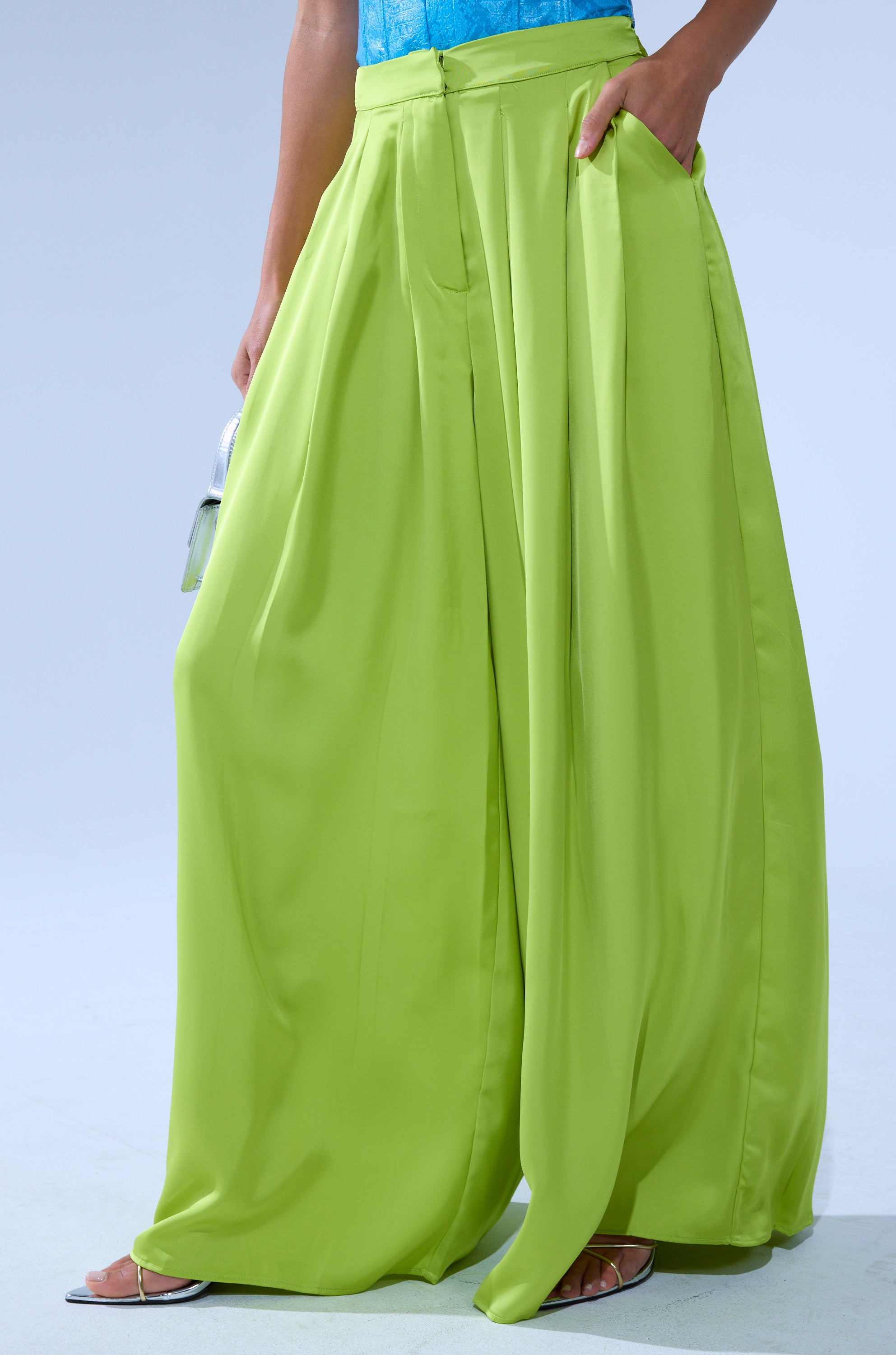 Wearing CHARTREUSE PLEATED PALAZZO PANT with wide legs, paired with an aqua blue top. One hand is in a pocket, while the other holds a small silver clutch. Only the lower half is shown. 3