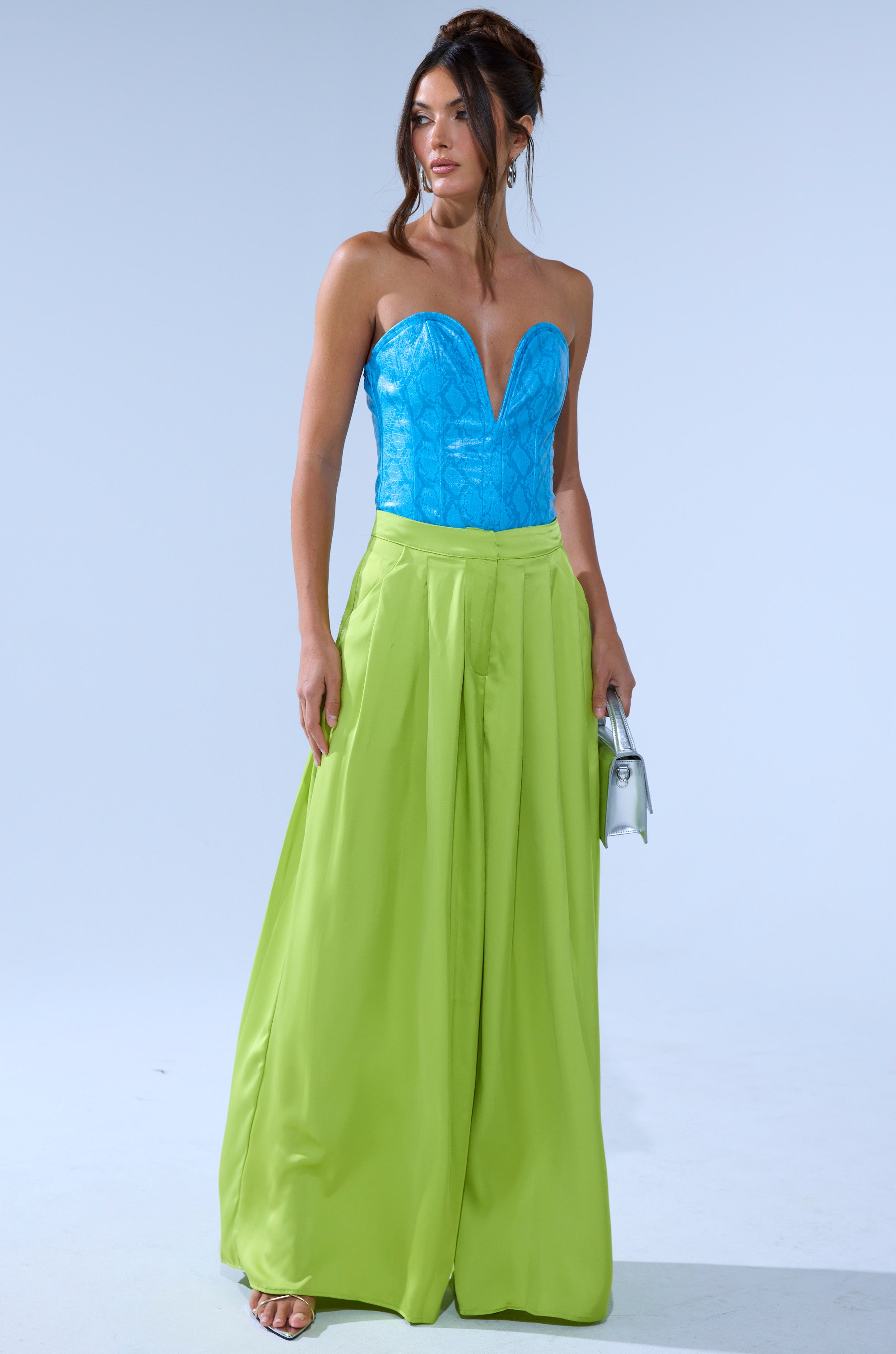 A woman stands against a plain light background wearing the CHARTREUSE PLEATED PALAZZO PANT with wide legs, paired with a blue strapless corset top. She holds a silver clutch, wears strappy heels, and her hair is styled up. 5
